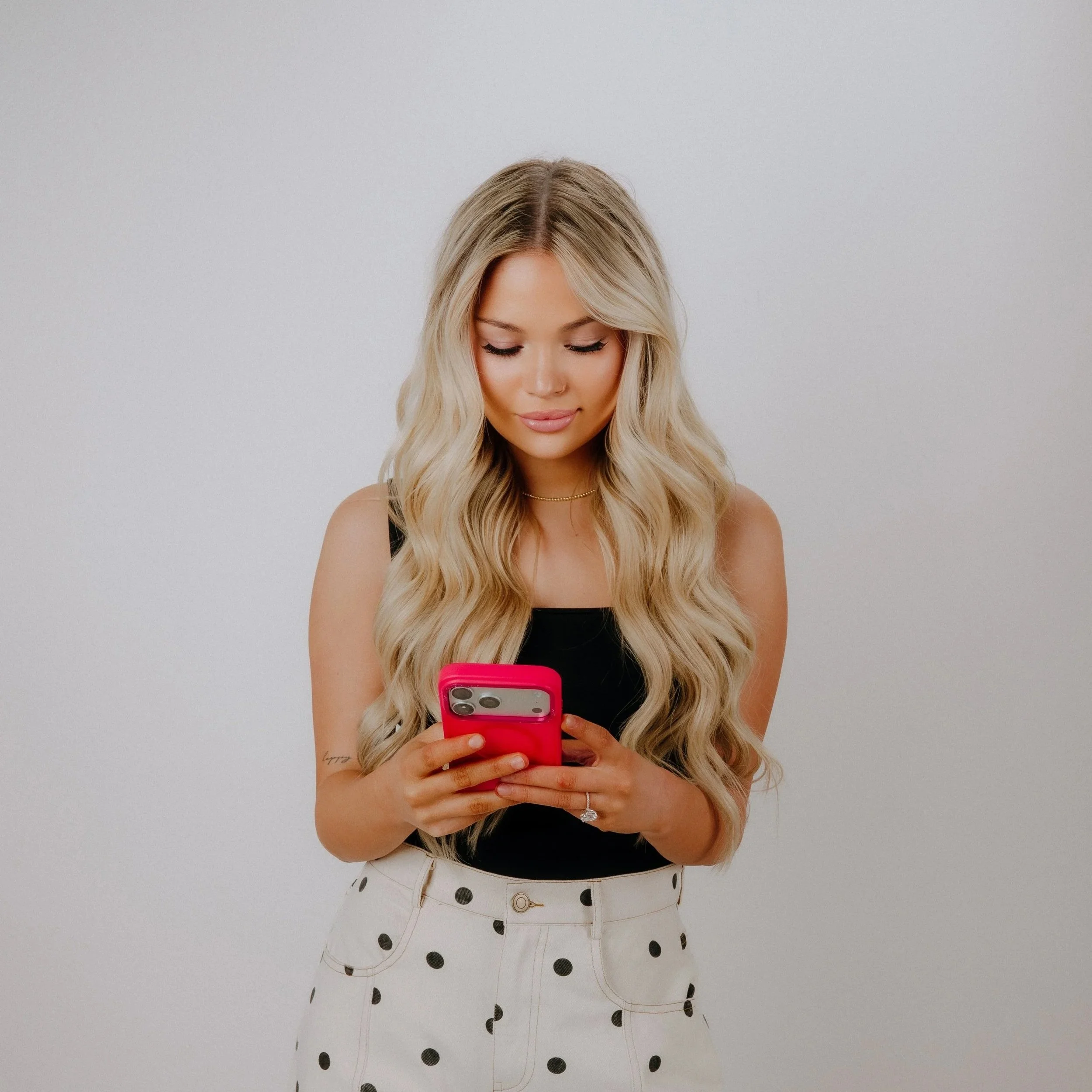 A woman with long wavy blonde hair, wearing a black sleeveless top and white skirt with black polka dots, looking down at her pink mobile phone against a plain background.