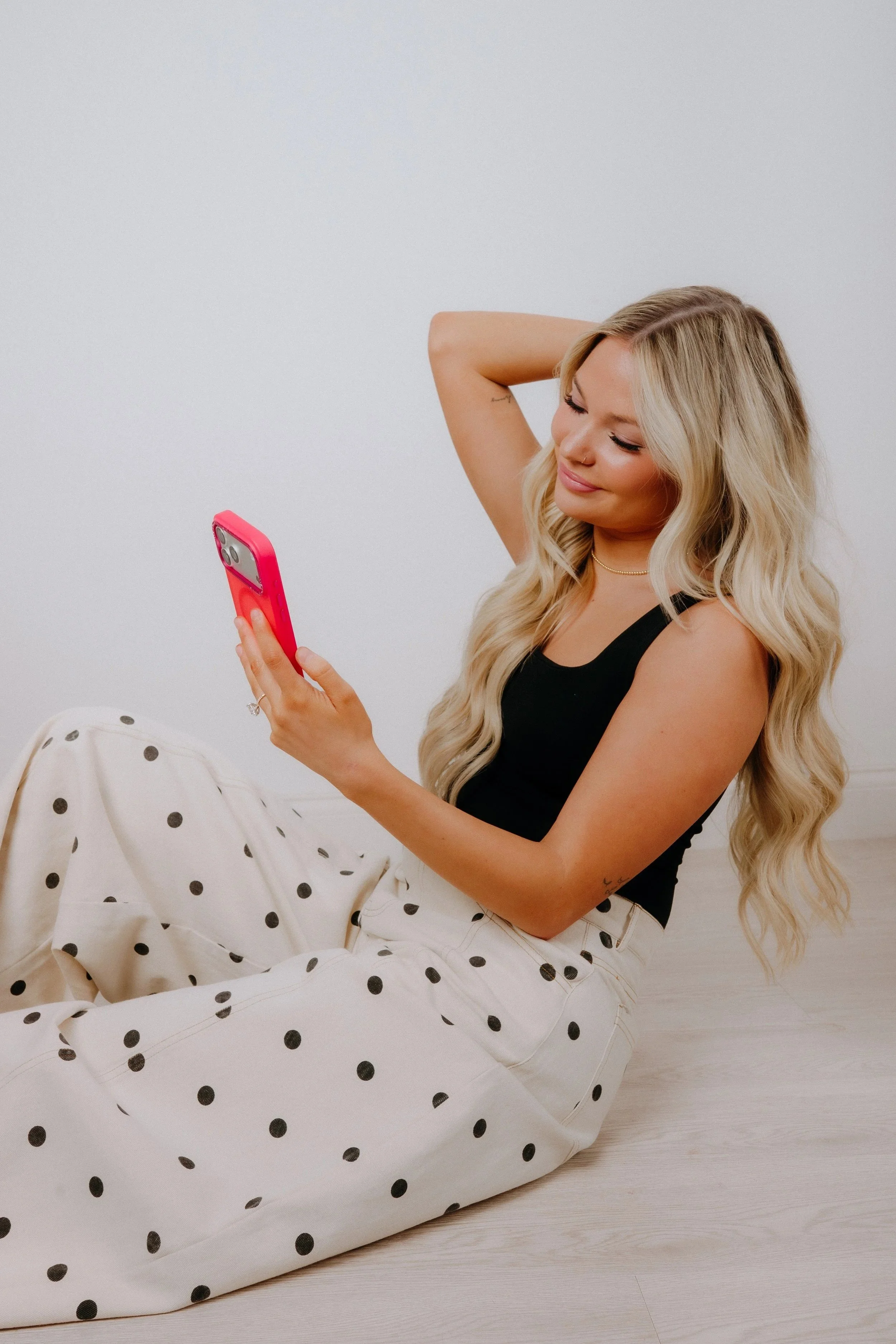 A young woman with long blonde wavy hair, wearing a black sleeveless top and white polka-dot pants, sitting on a light-colored wooden floor, holding a pink phone and smiling.