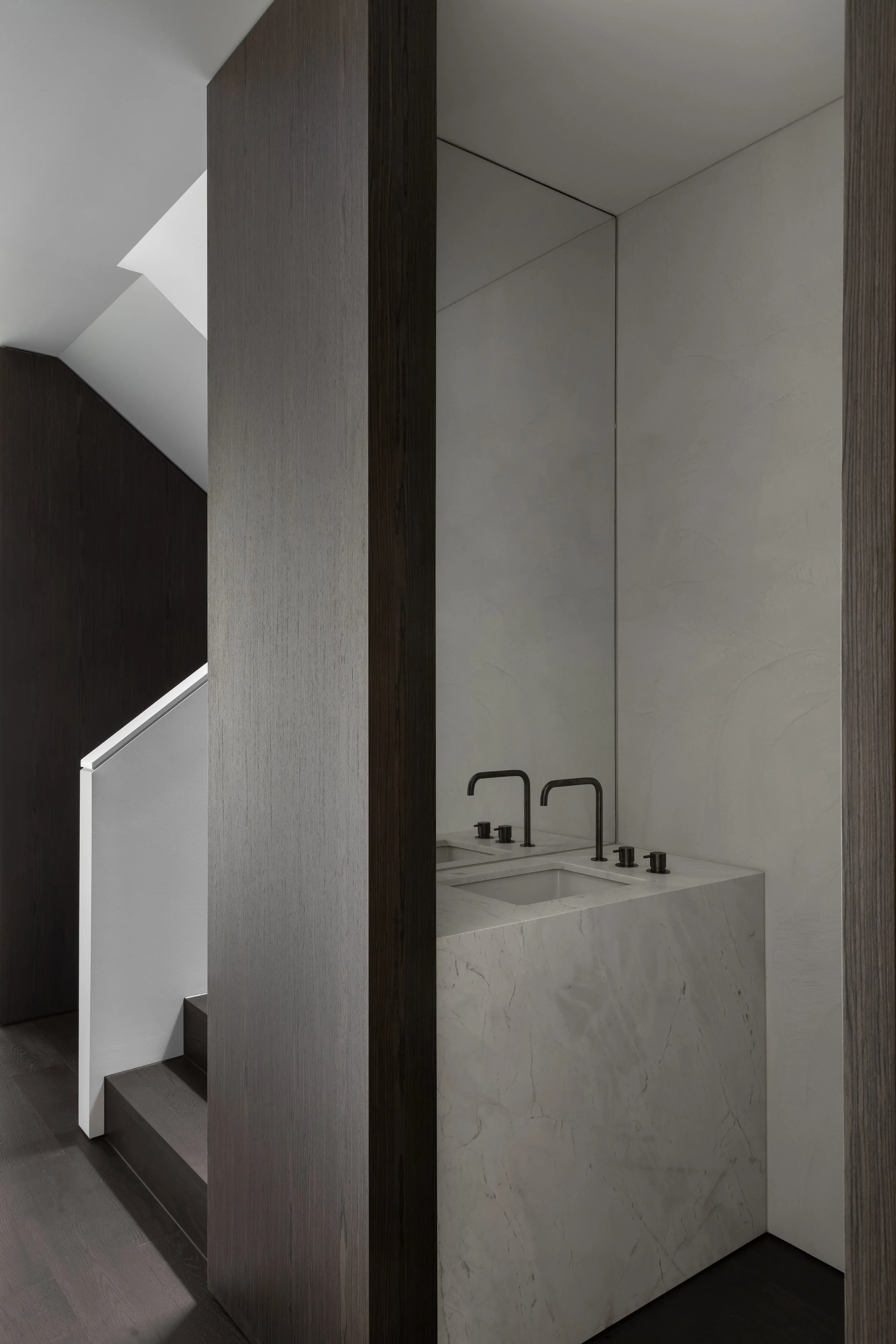 A modern bathroom sink with two black faucets and black knobs set into a white marble countertop, behind a partial wall with dark wood paneling, next to a staircase.