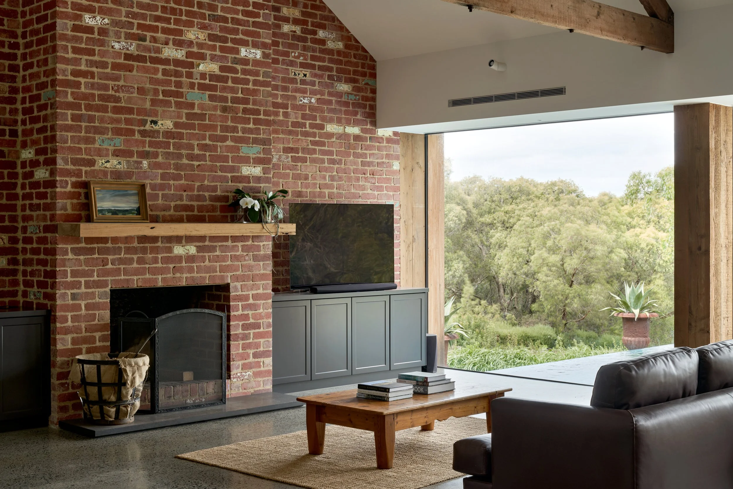 Living room with brick fireplace, gray media cabinet, flat-screen TV, wooden coffee table, black leather sofa, large window with view of outdoor greenery, potted plants, and ceiling beam