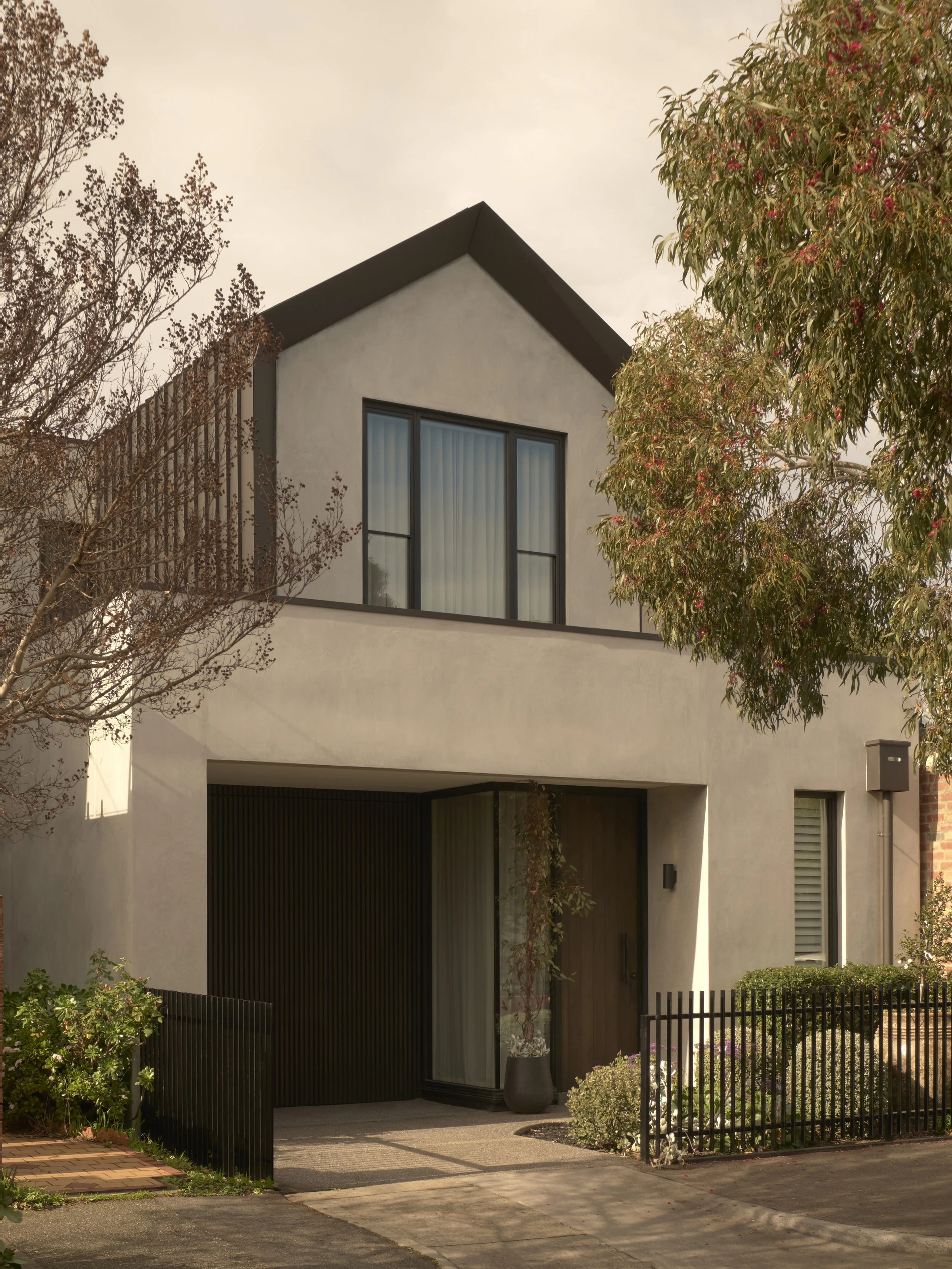 Modern two-story house with white exterior, large front window, and black metal fence, surrounded by trees and bushes.