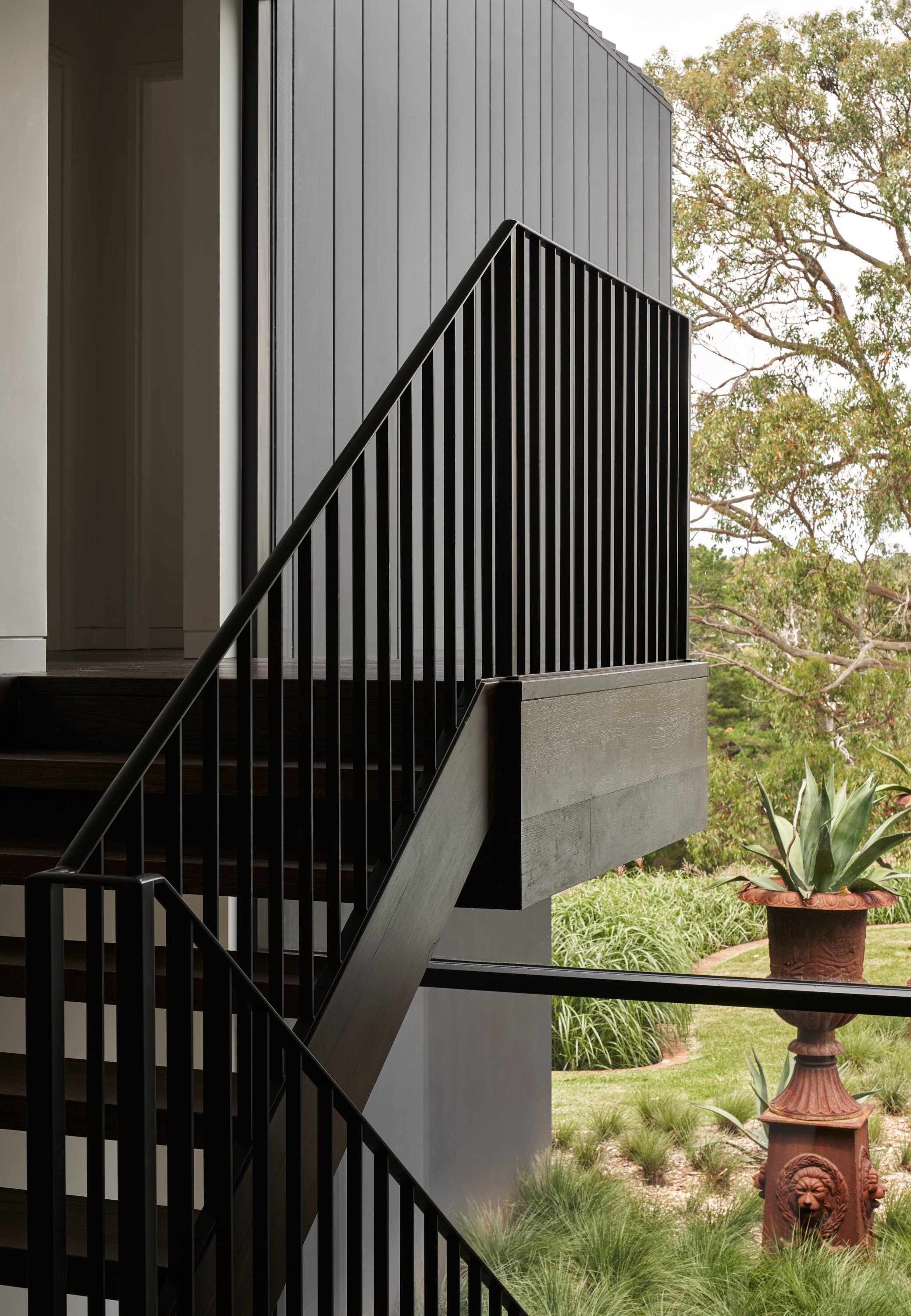 Exterior view of a modern building with black metal staircase railing, black siding, and a potted plant outside with trees and grass in the background.