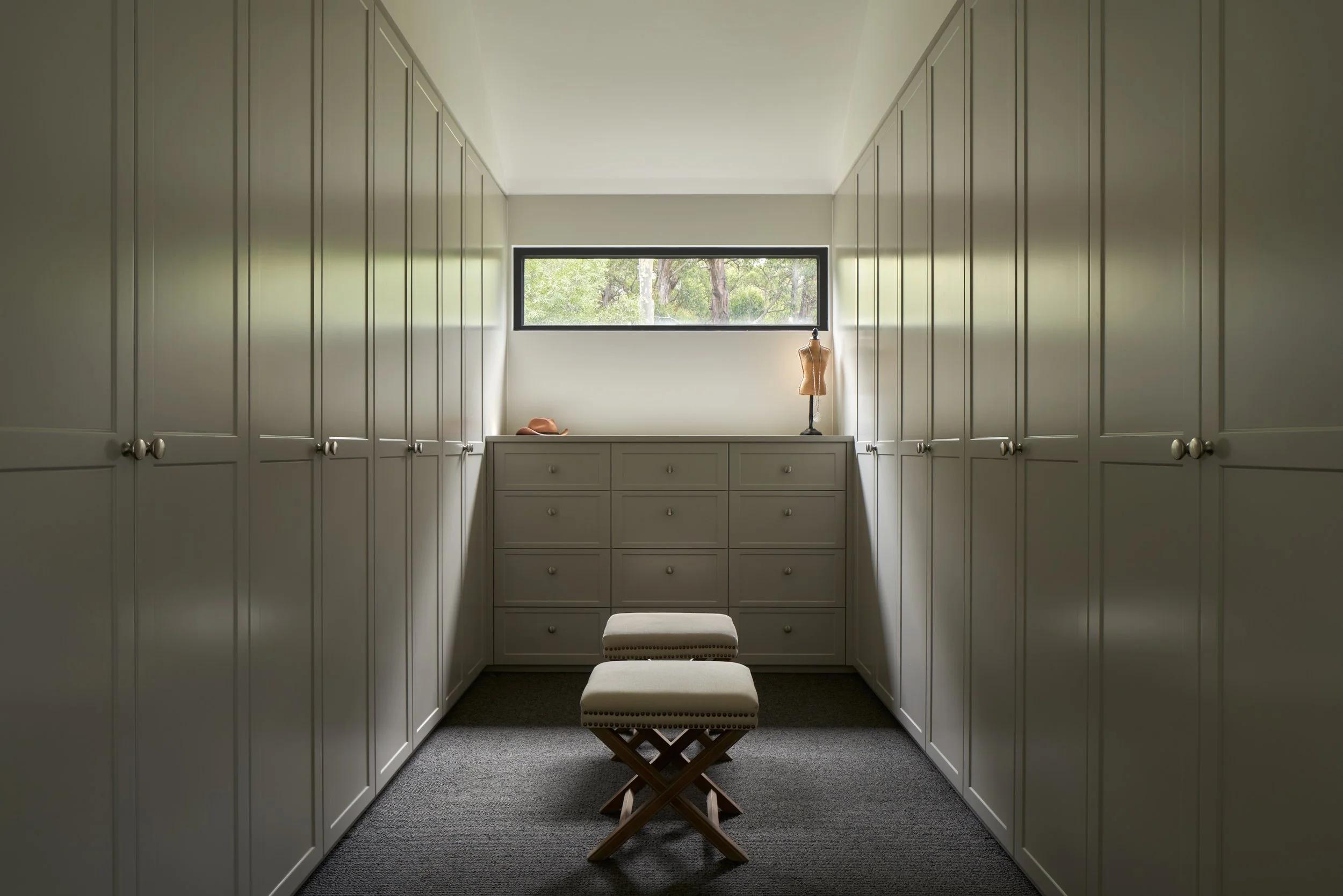A walk-in closet with white cabinets on both sides, a window at the end, and a white dresser with a dress form, hat, and other items on top. Two upholstered stools are in the center.