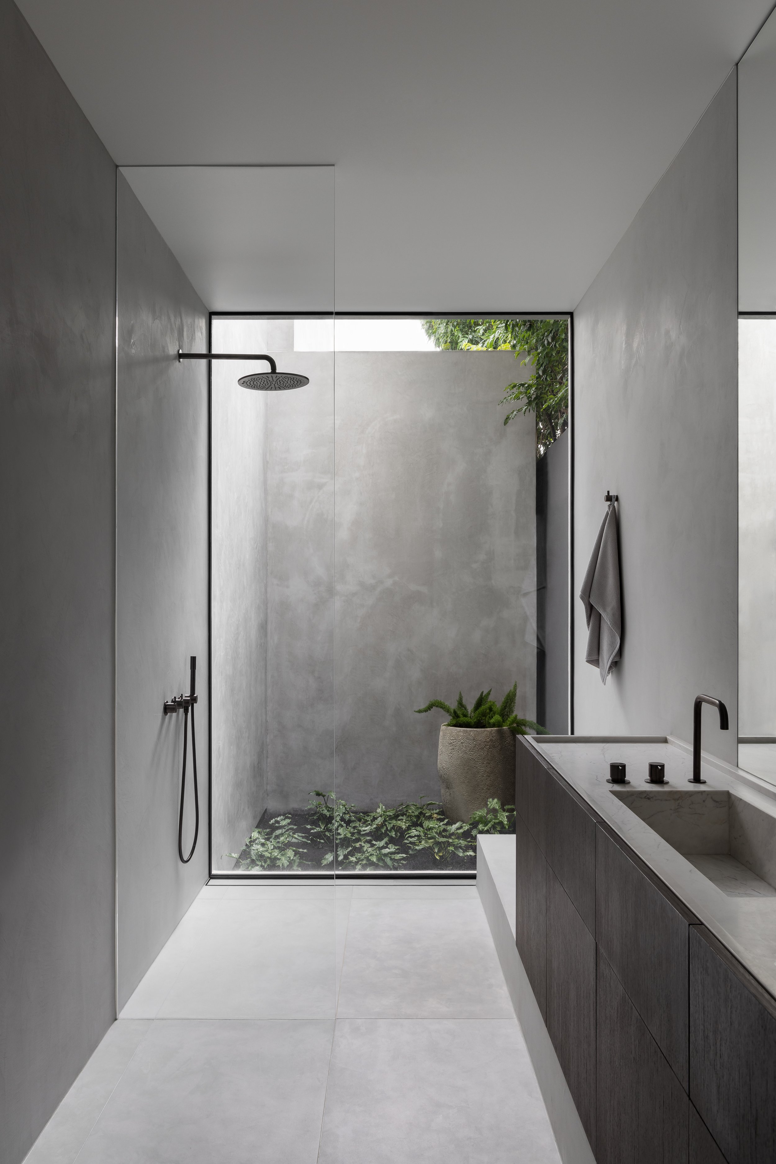 Modern minimalist bathroom with walk-in shower with glass wall, rain showerhead, grey wall and floor tiles, a dark vanity with a sink, a towel hanging on the wall, and a large window overlooking a small plant garden.