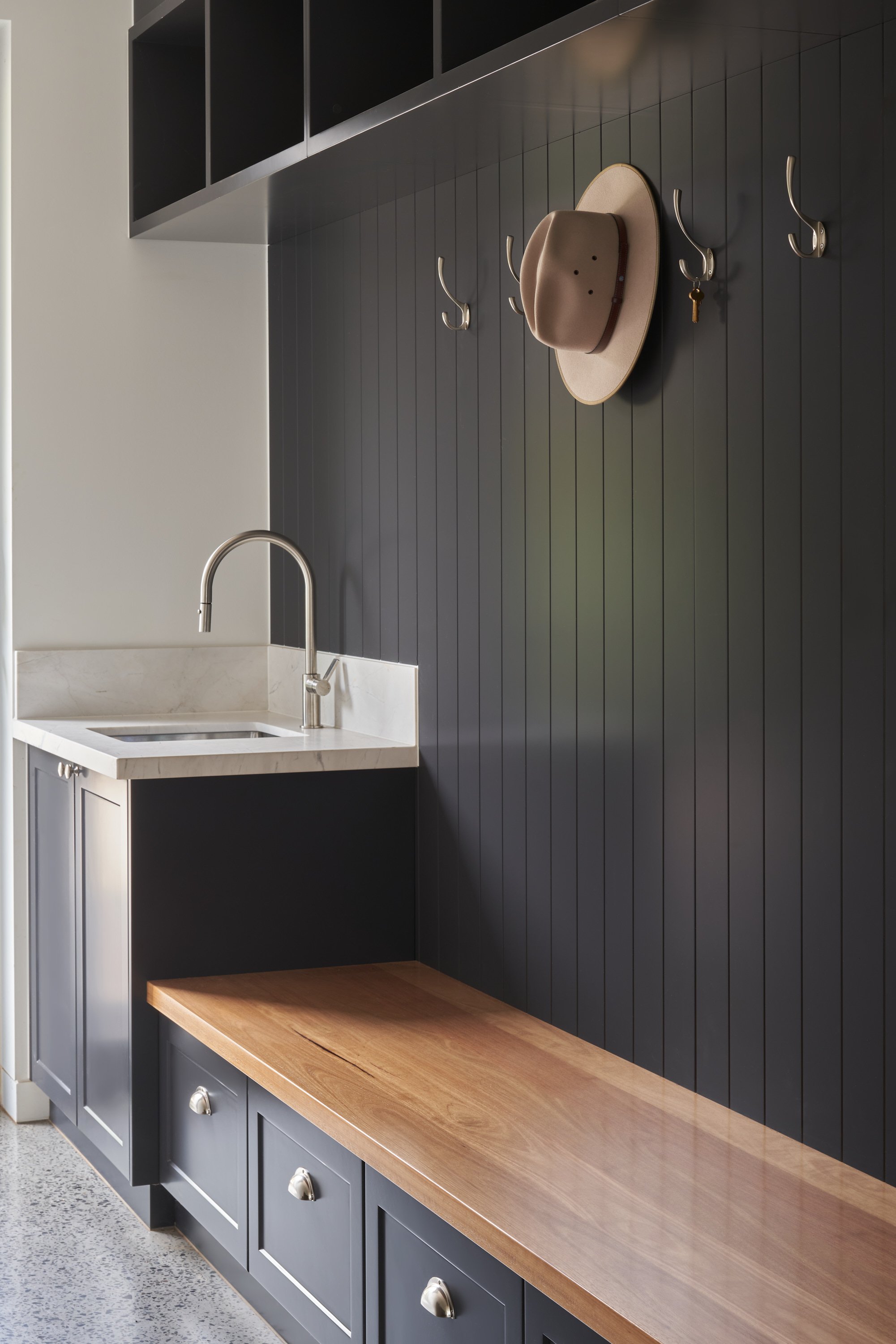 A modern mudroom with dark paneled wall, hooks holding a hat and keys, a small sink with a faucet, and a wooden bench with storage drawers underneath.