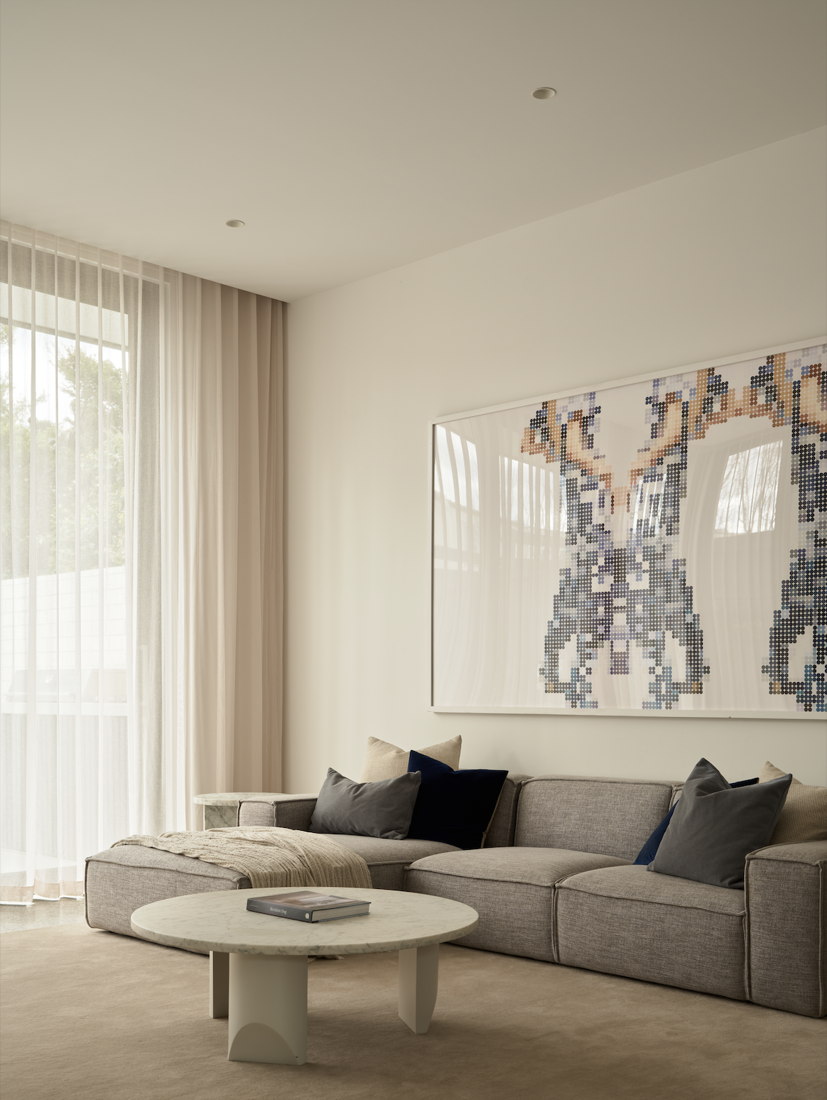 Living room with large window, beige curtains, gray sofa with multiple pillows, a cream blanket, a round marble coffee table with a book, and a pixelated artwork of a butterfly on the wall.