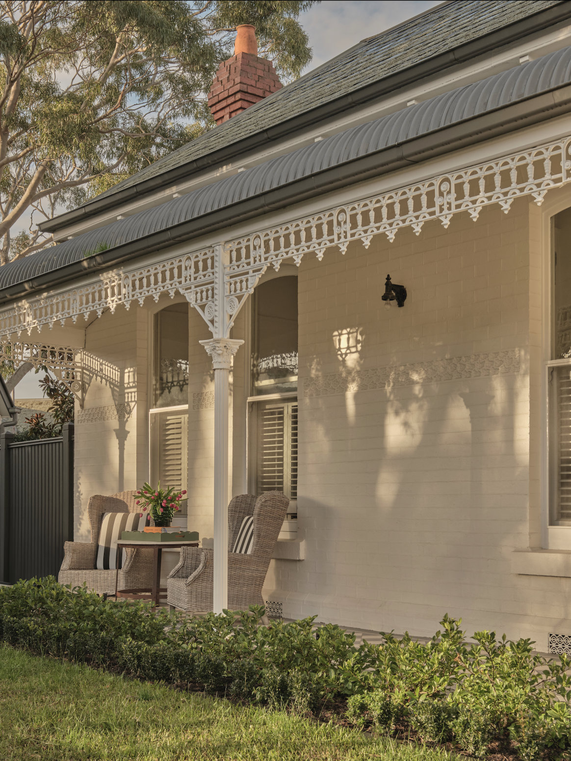 Front porch of a house with a white brick facade, ornate white trim, wicker chairs, a small wooden table with a flower arrangement, and lush greenery in the foreground.