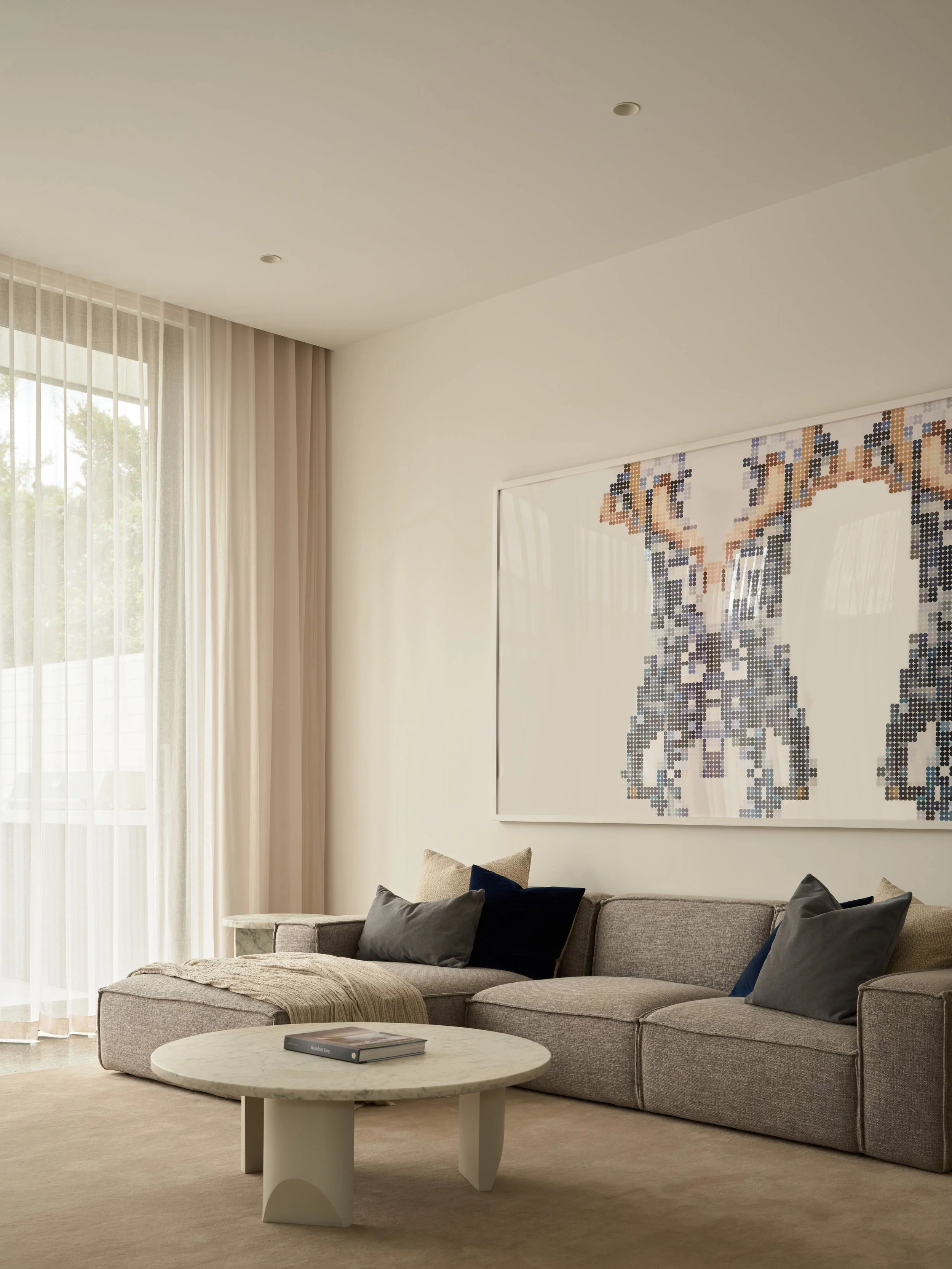 Modern living room with beige sofa, multicolored pillows, a round marble coffee table, sheer curtains, and a large pixelated artwork of a dog on the wall.