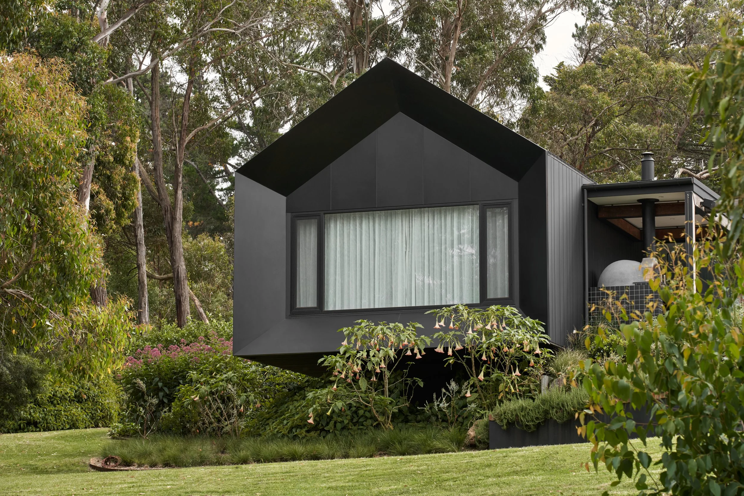Modern black house with large window, surrounded by greenery and trees.
