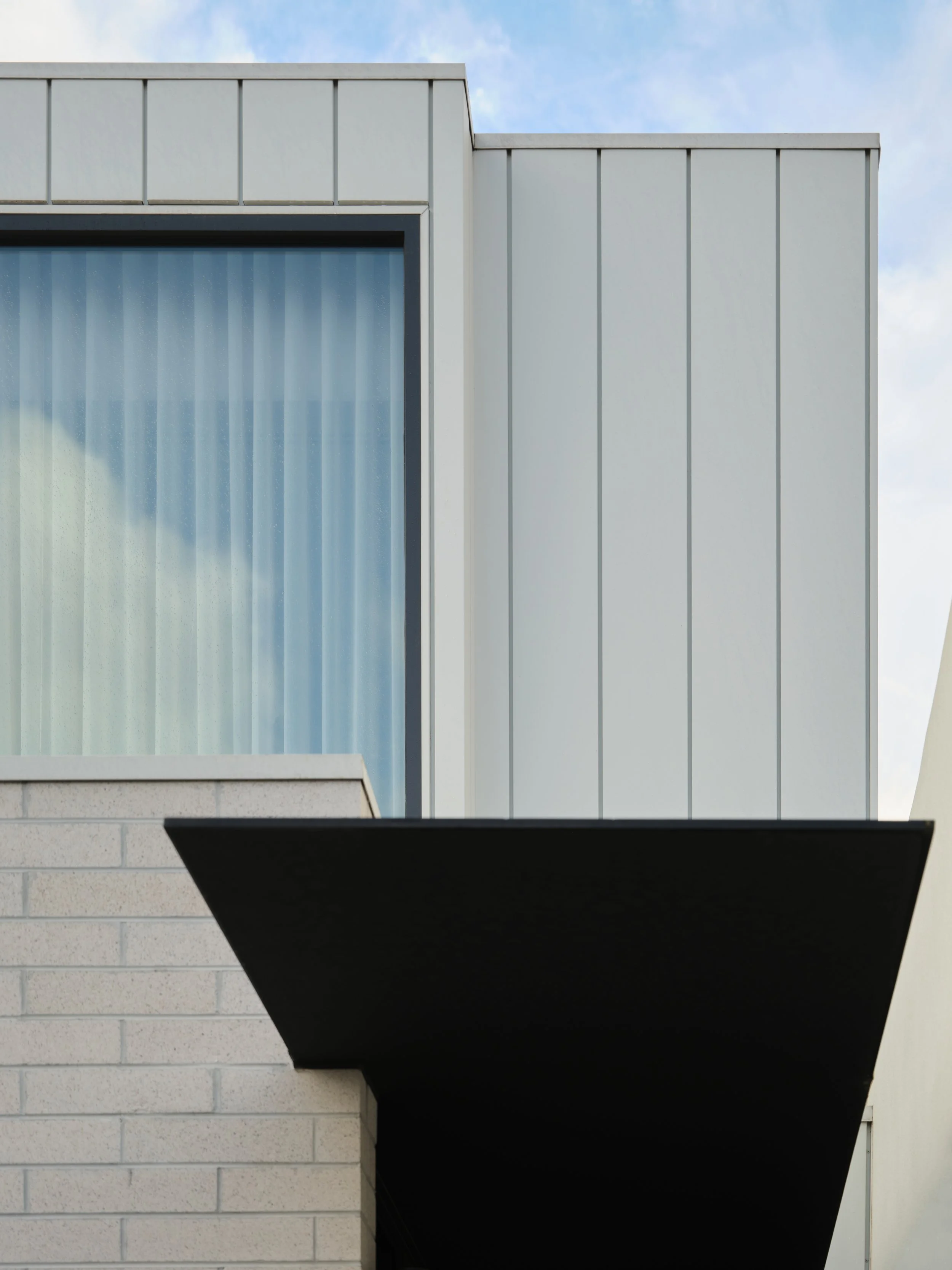 Close-up of a modern building exterior with a large window, white panel siding, and a black awning against a blue sky with clouds.
