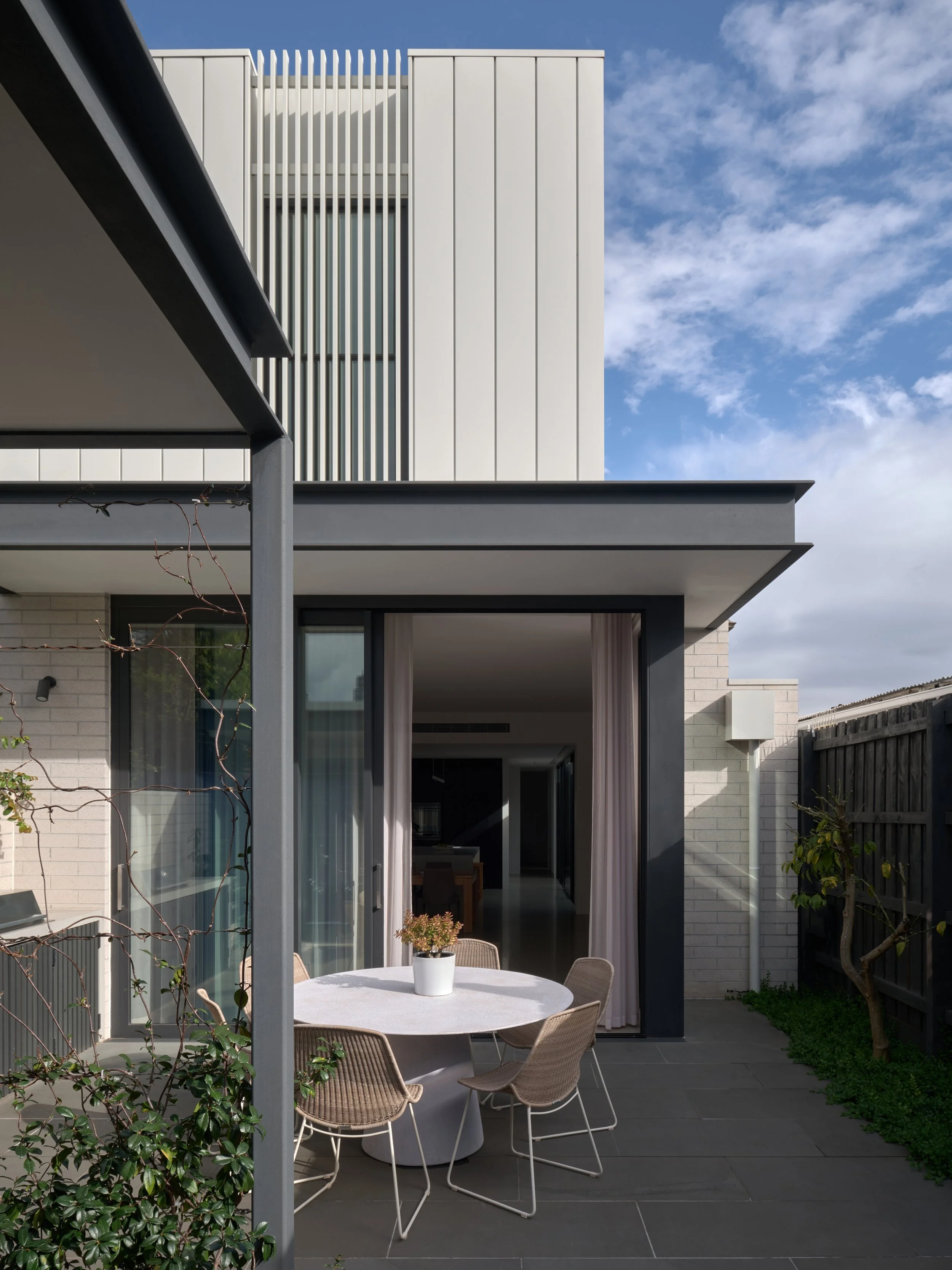 Modern house patio with round table, four chairs, potted plant, and sliding glass door, under a clear sky.