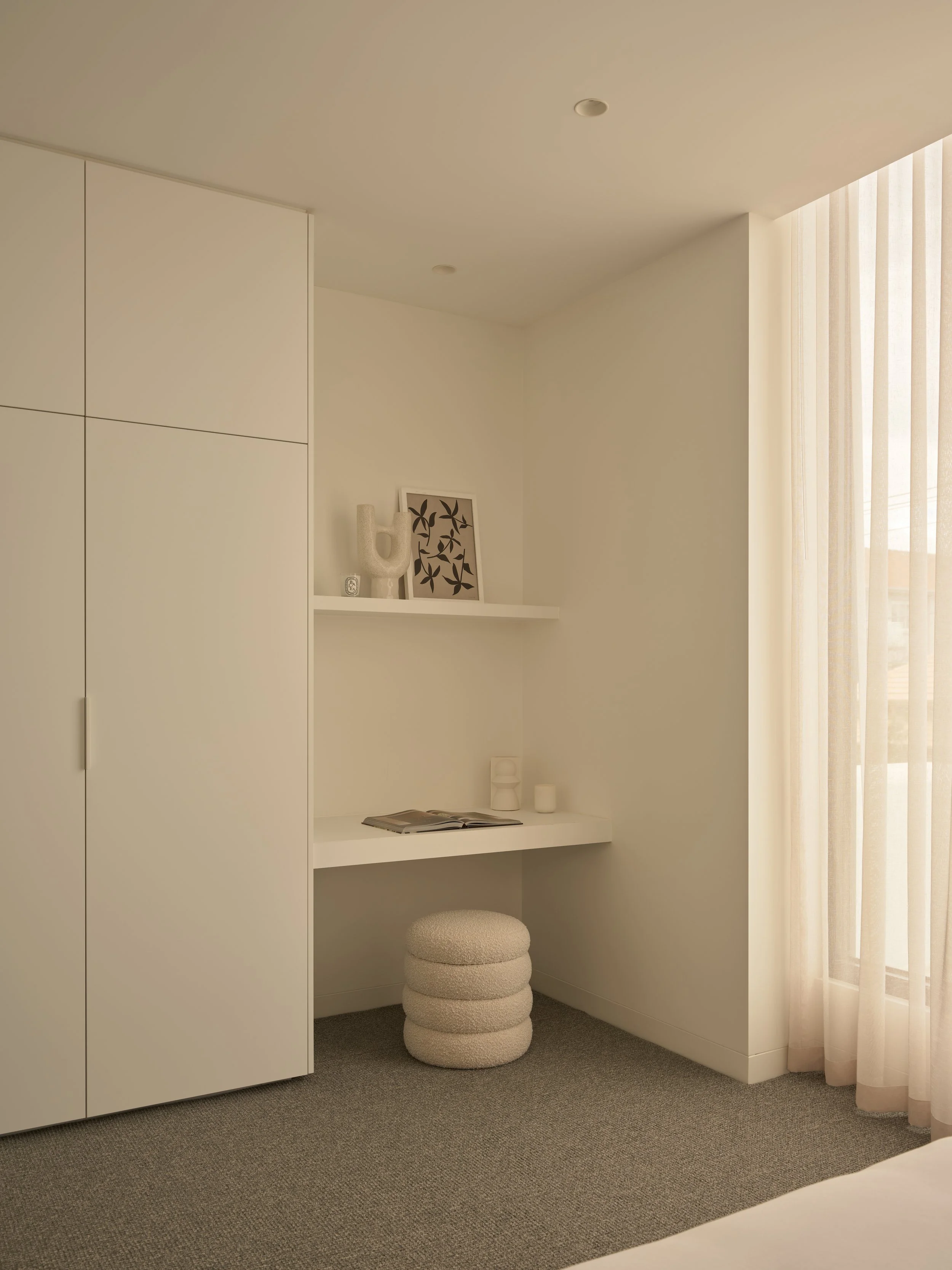 Minimalist bedroom corner with white built-in wardrobe, small white desk, and beige pouf stool, with decor objects and sheer curtains.