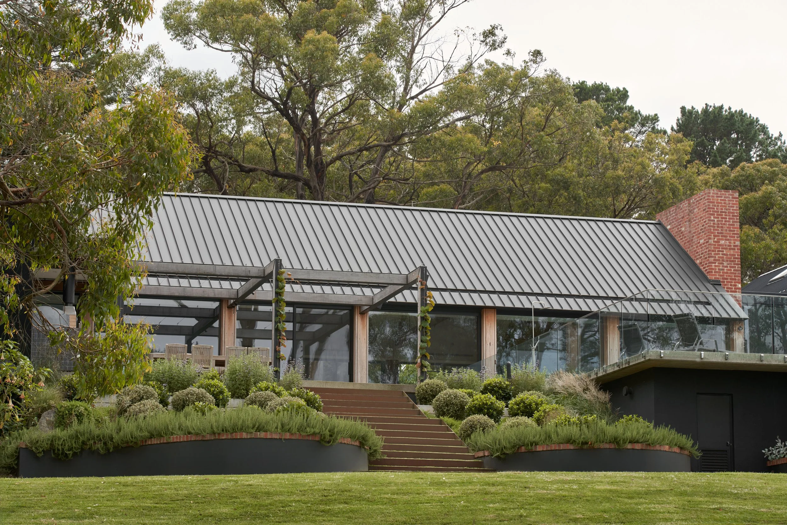 A modern house with a metal roof, large glass windows, and a chimney, surrounded by landscaped gardens with bushes, stairs leading up to the house, and trees in the background.