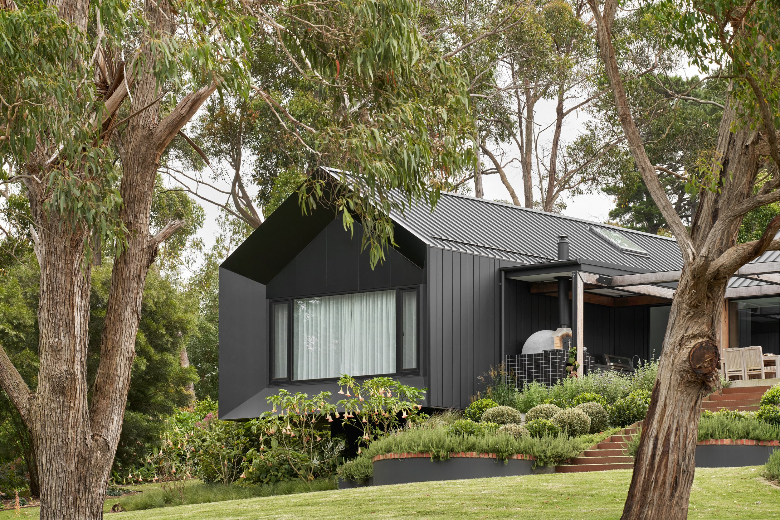 A modern black house with a slanted roof, large windows with white curtains, in a lush green yard with tall trees and landscaped plants.