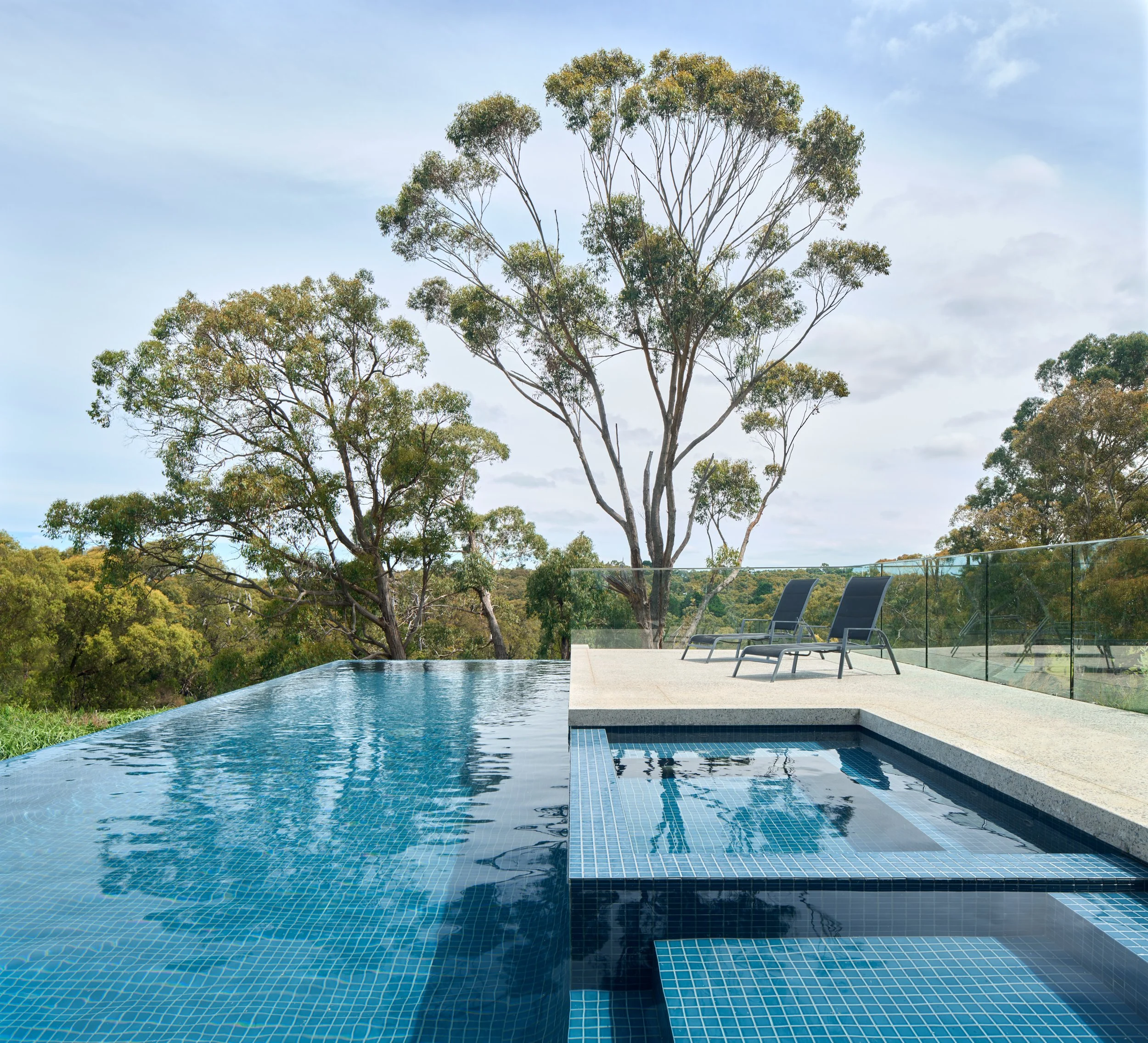 Infinity pool with blue tiles overlooking trees and a cloudy sky, with two black lounge chairs on a patio.