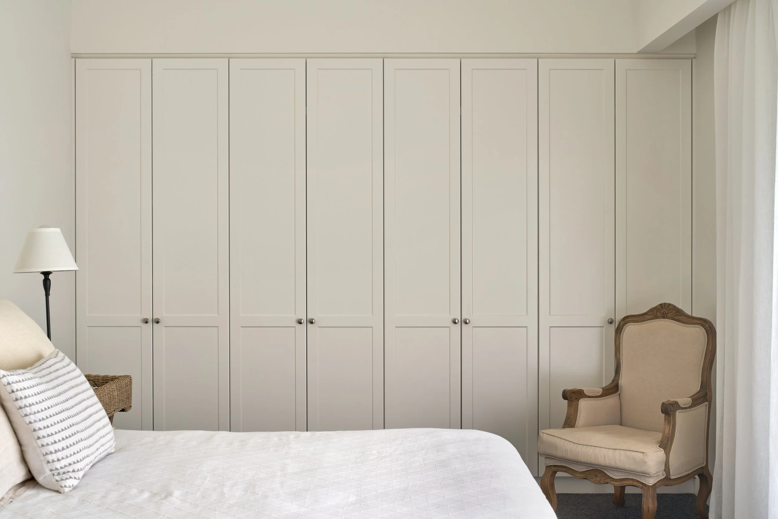 A bedroom with a white wardrobe, a bed with white linens and striped pillows, a white bedside lamp, and an antique armchair.