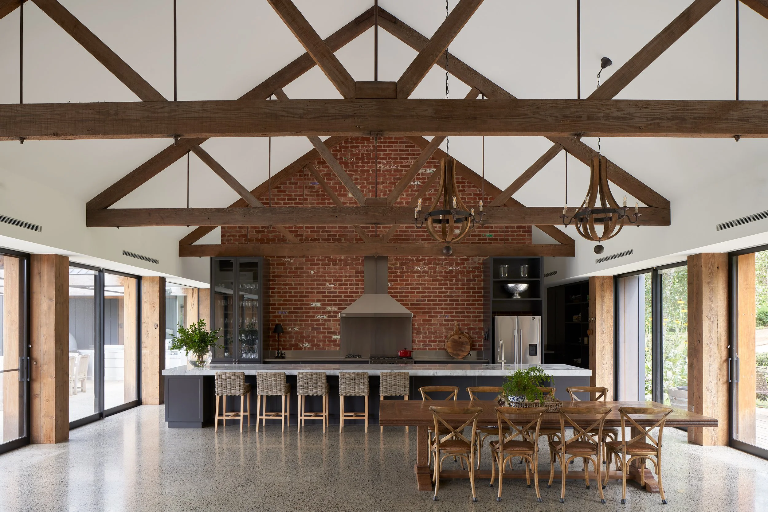Modern kitchen and dining area with a brick wall, wooden beams, chandelier, large windows, black cabinetry, and a kitchen island with barstools.