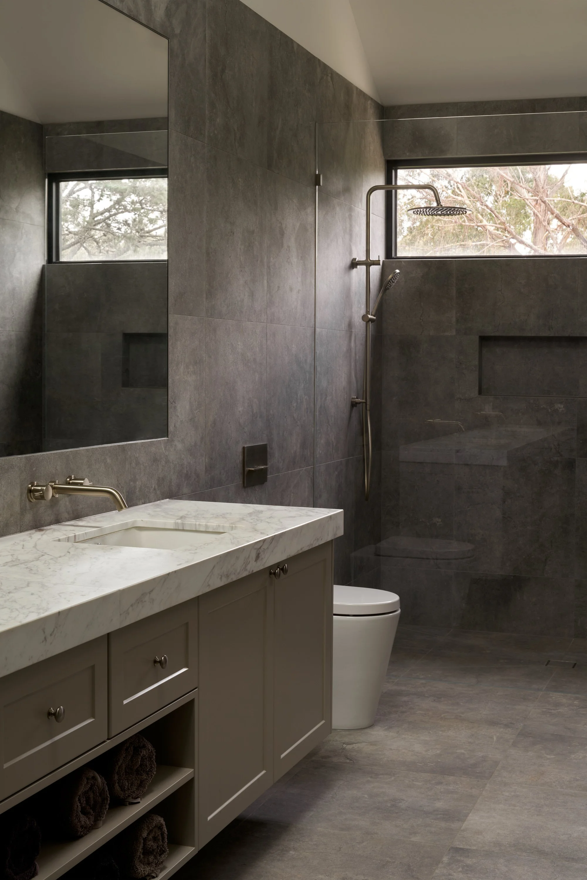 Modern bathroom with dark gray tiled walls and floor, a white vanity with a marble top, a sconce, a toilet, and a walk-in shower with a window, rain showerhead, and a handheld shower.