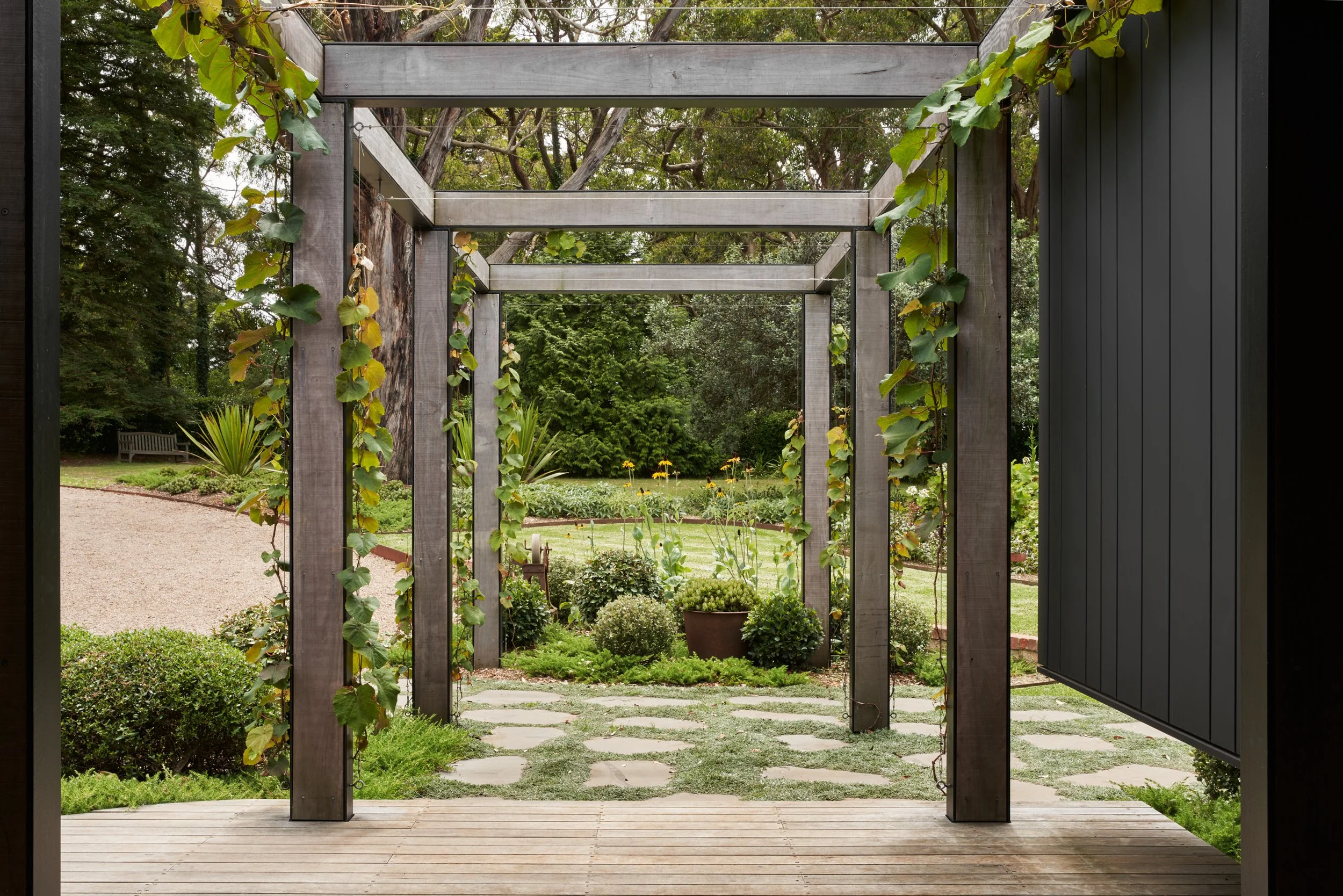 View from a covered porch with wooden decking, looking out onto a garden with a trellis, stepping stones, various plants, and trees in the background.