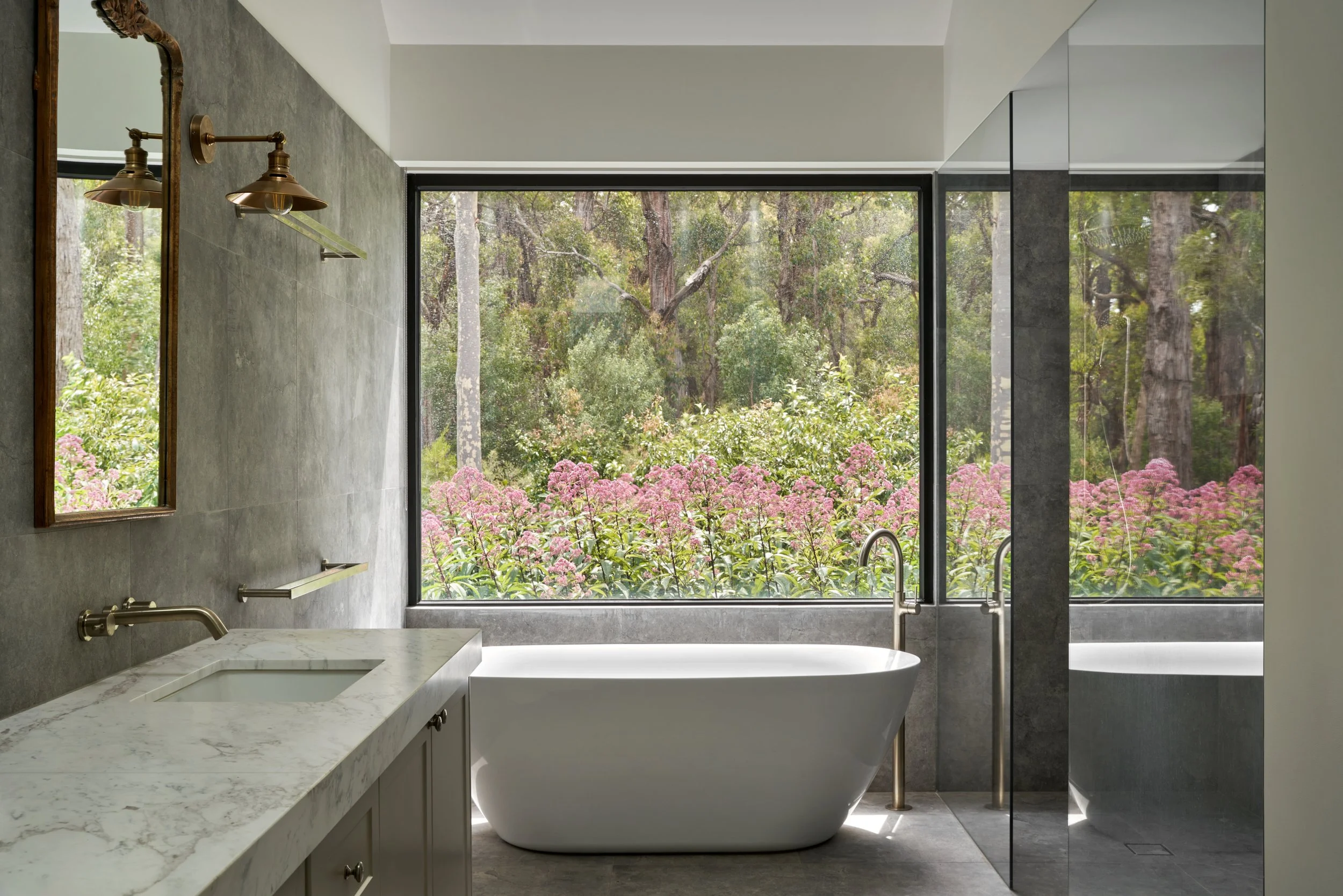 Modern bathroom with large window showing garden with pink flowers and trees outside; features a white bathtub, marble countertop, mirror, and brass fixtures.