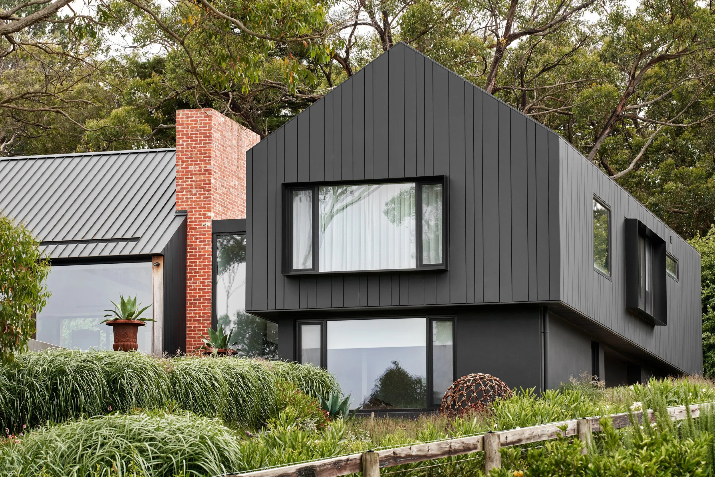 Modern black house with large windows and a red brick chimney, surrounded by green plants and trees.