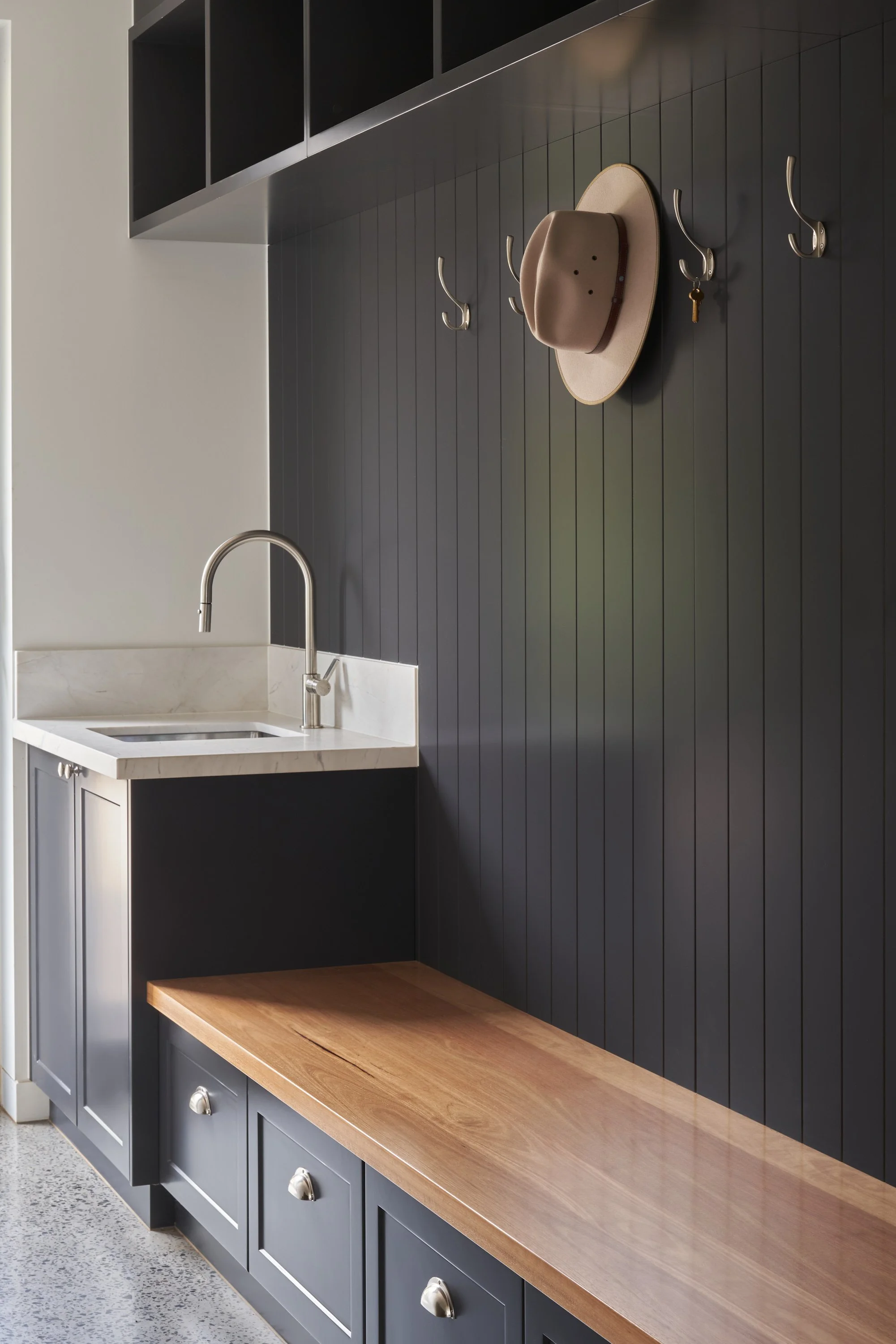 A modern entryway with black panel wall, hooks, a hat, and a key. A small sink with a silver faucet and large cabinet with drawers below a wooden bench are visible.