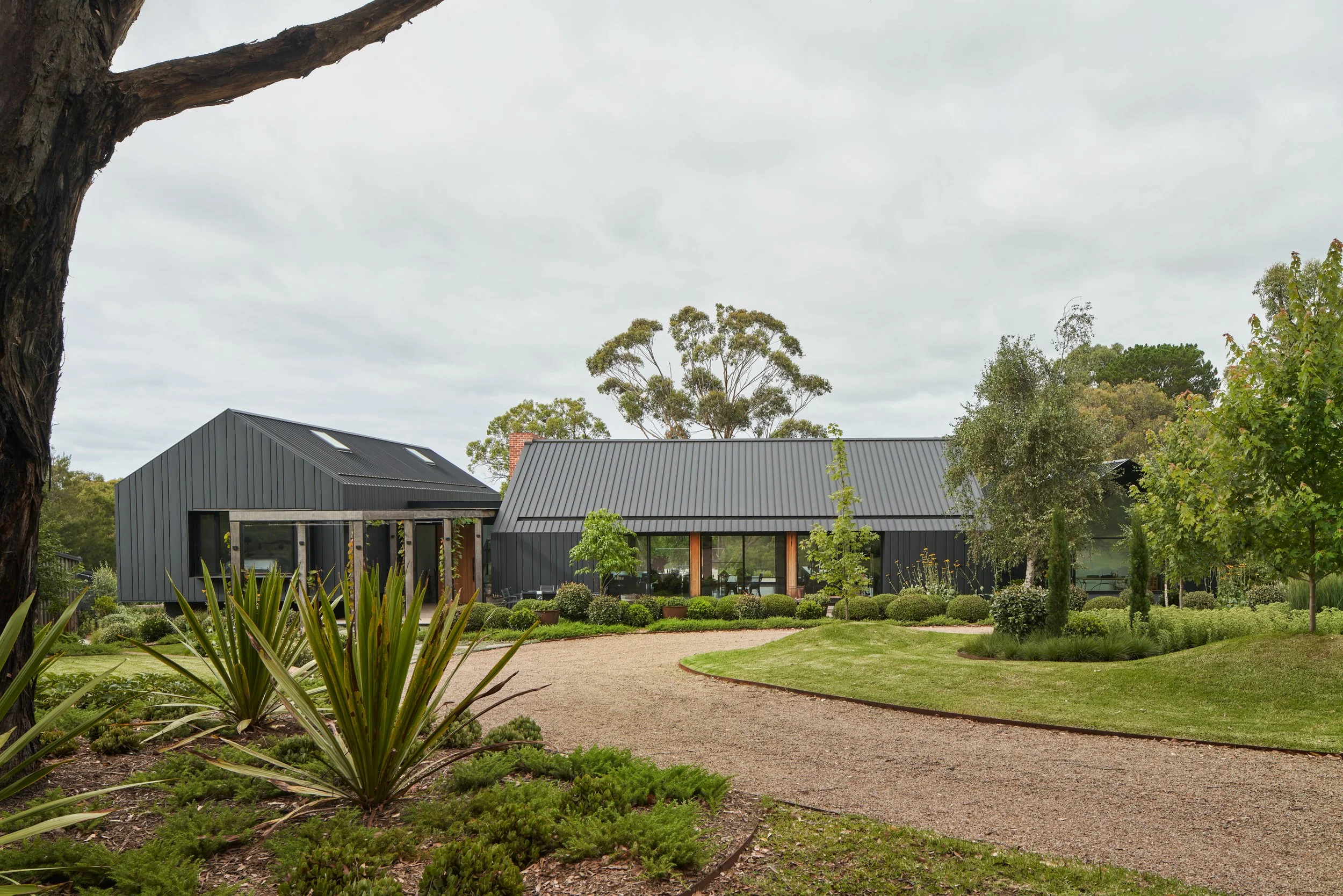 Modern house with black metal siding and large glass windows, surrounded by a lush green garden and trees, with a gravel pathway in the foreground.