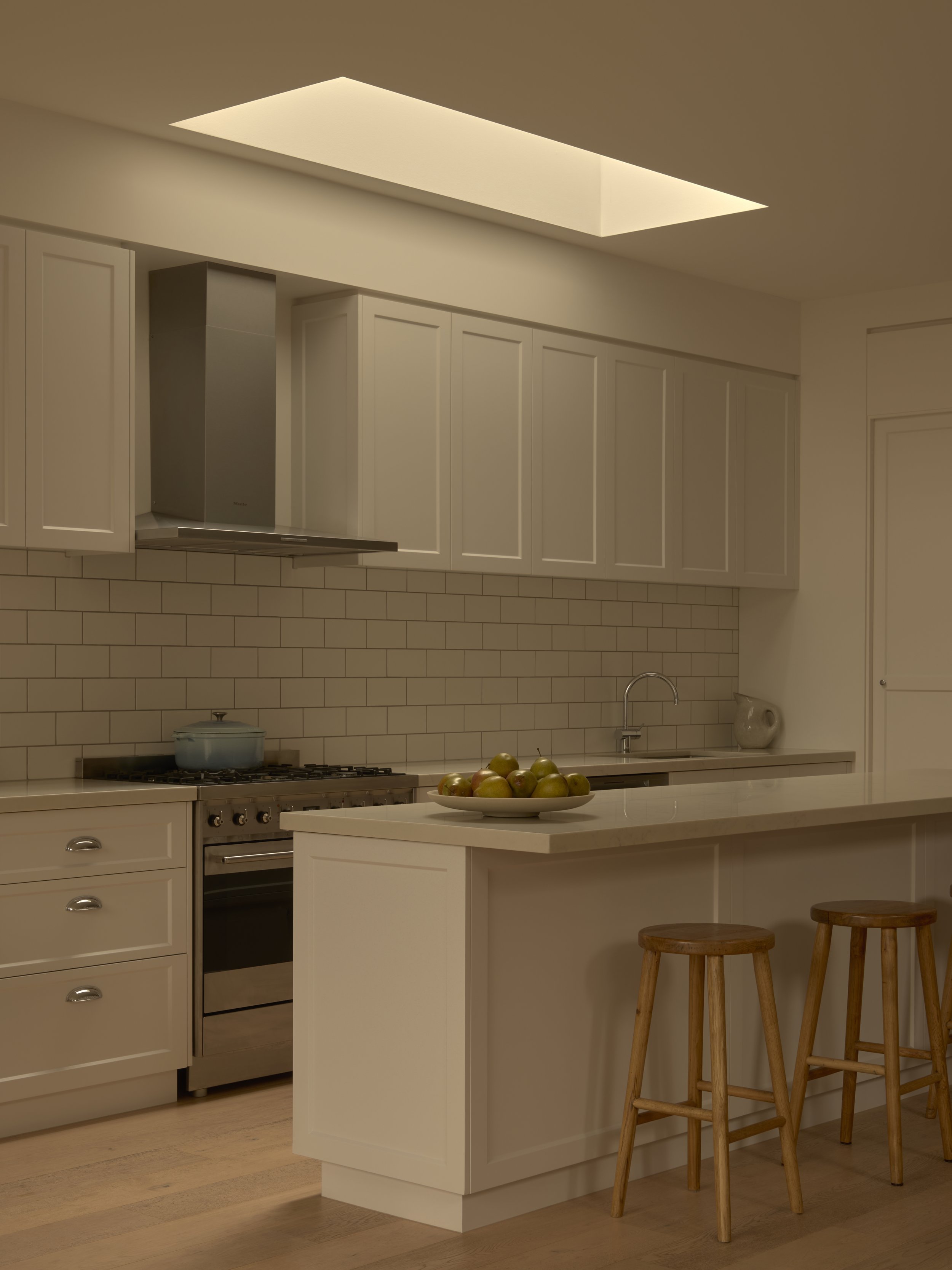 Modern kitchen with beige cabinets, white island with wooden stools, stove with pot, and a bowl of green apples.