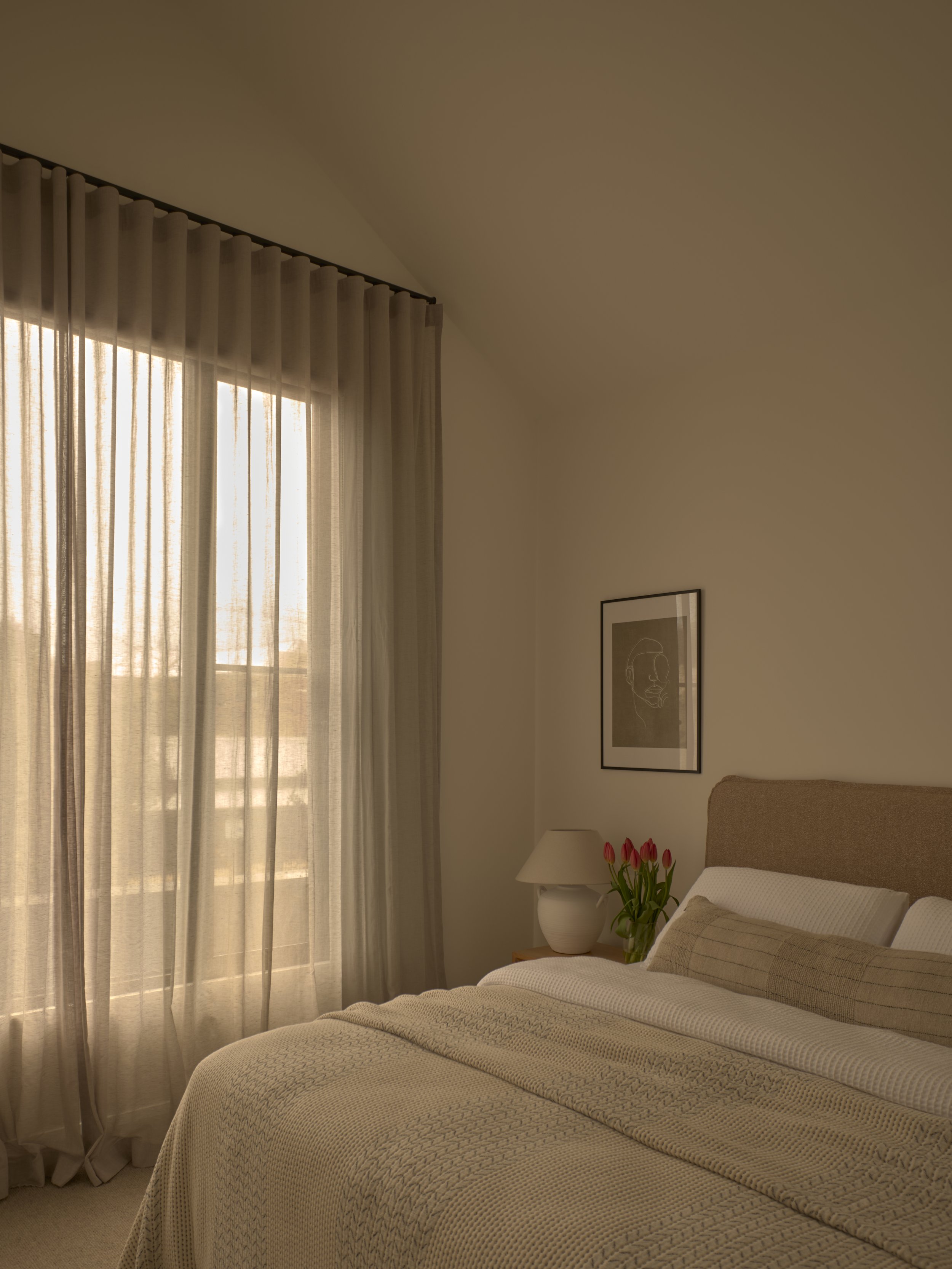 A cozy bedroom with beige bedding, a headboard, a nightstand with a lamp, a vase of red tulips, framed artwork, and a window with curtains.