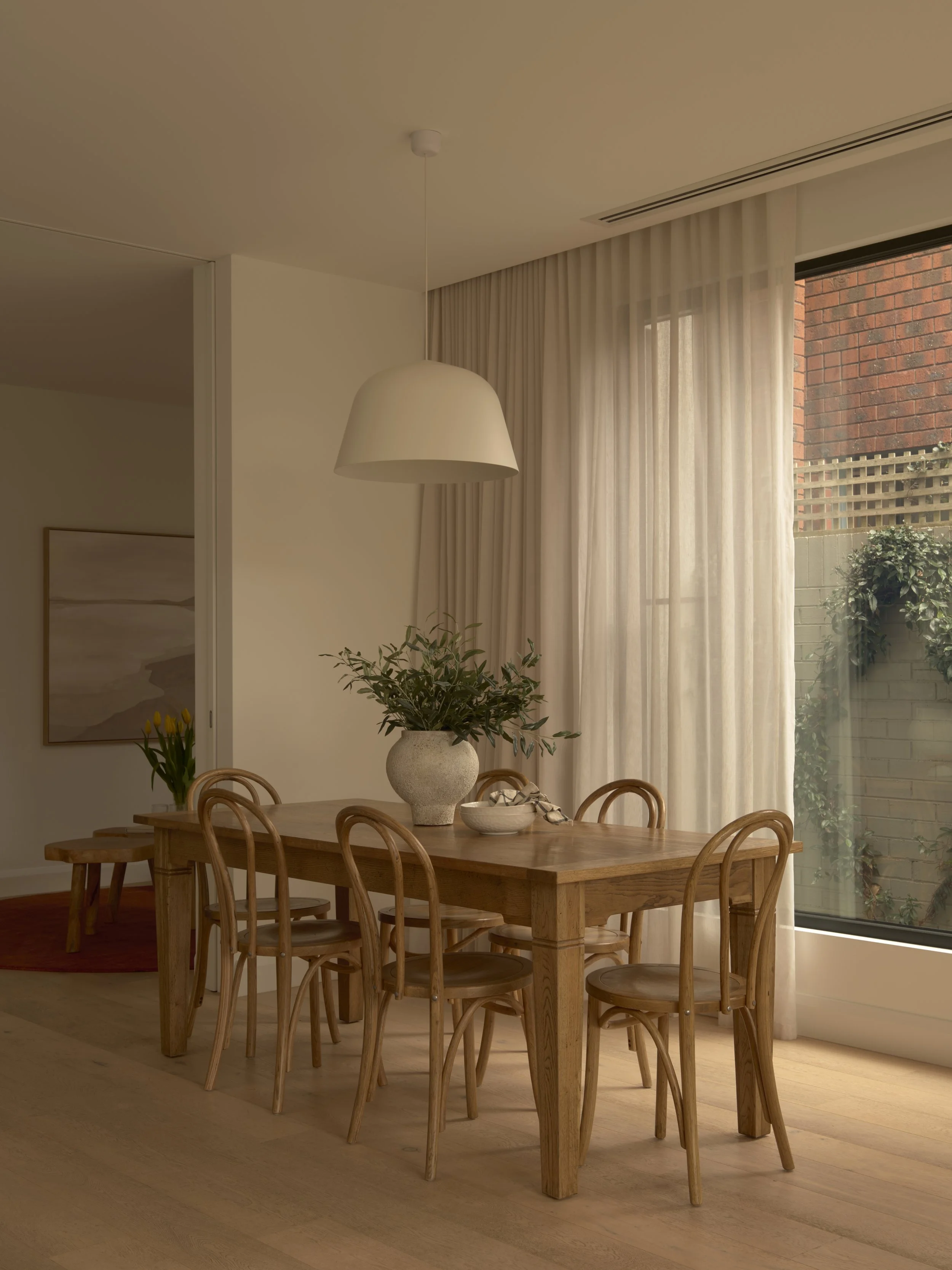 Modern dining room with a wooden table, six wooden chairs, a vase with greenery, and a large window with sheer curtains letting in natural light.