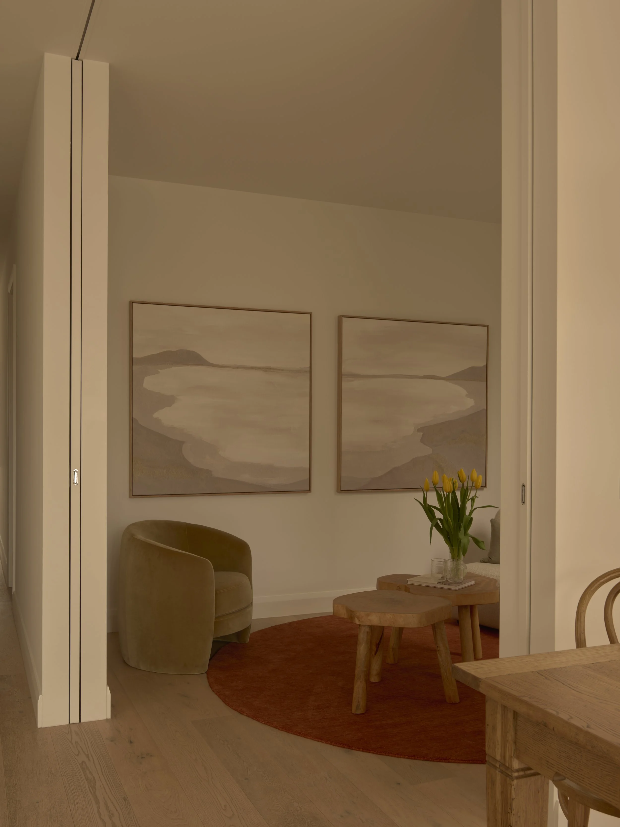 Cozy corner of a living room with a beige armchair, a round red rug, a wooden coffee table, yellow tulips in a glass vase, and two landscape paintings on the wall.