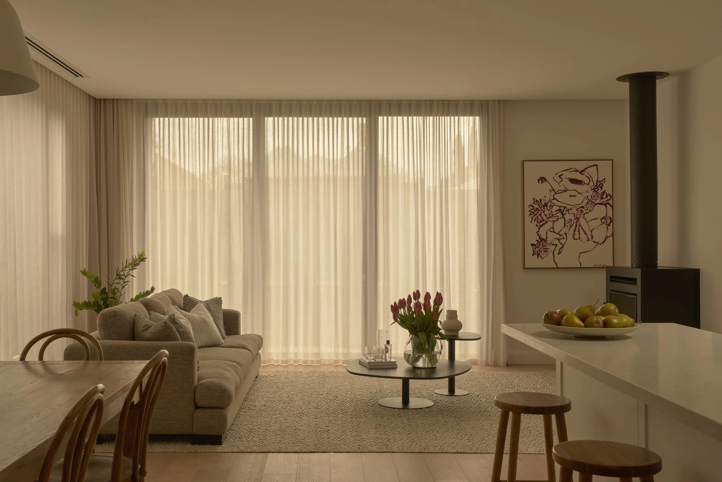 Bright living room with beige sofa, flower arrangement, and art on wall, lit by natural light through sheer curtains.