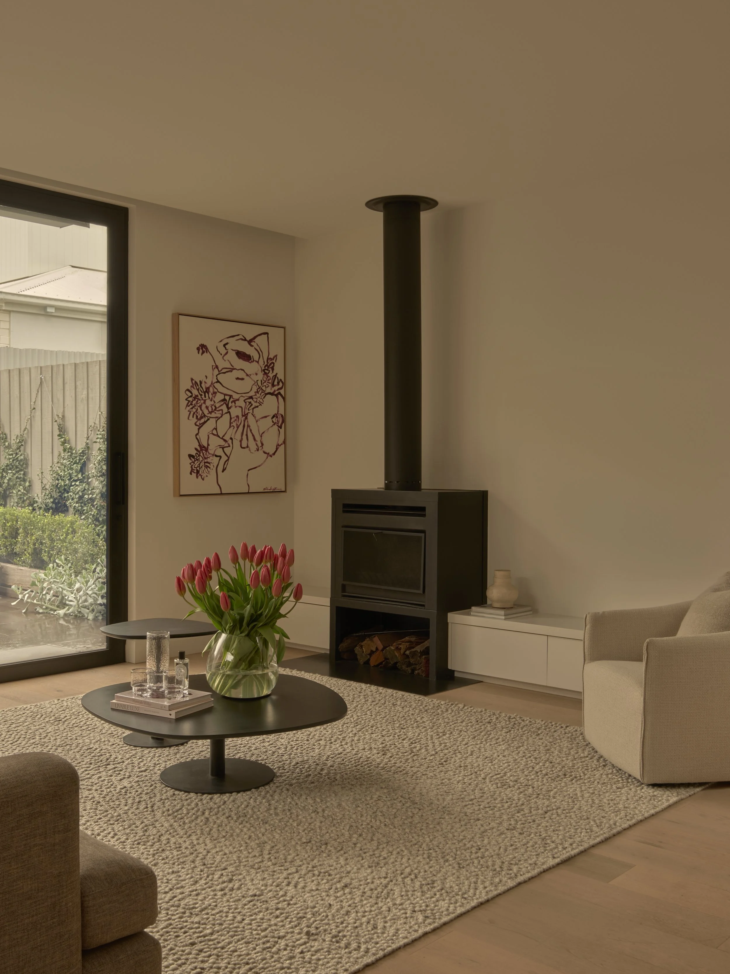 Living room with a beige armchair, a black round coffee table with a vase of pink tulips, a glass of water, and books, a fireplace with logs, a piece of abstract wall art, a sliding glass door shows a backyard with plants, and there's a white cabinet