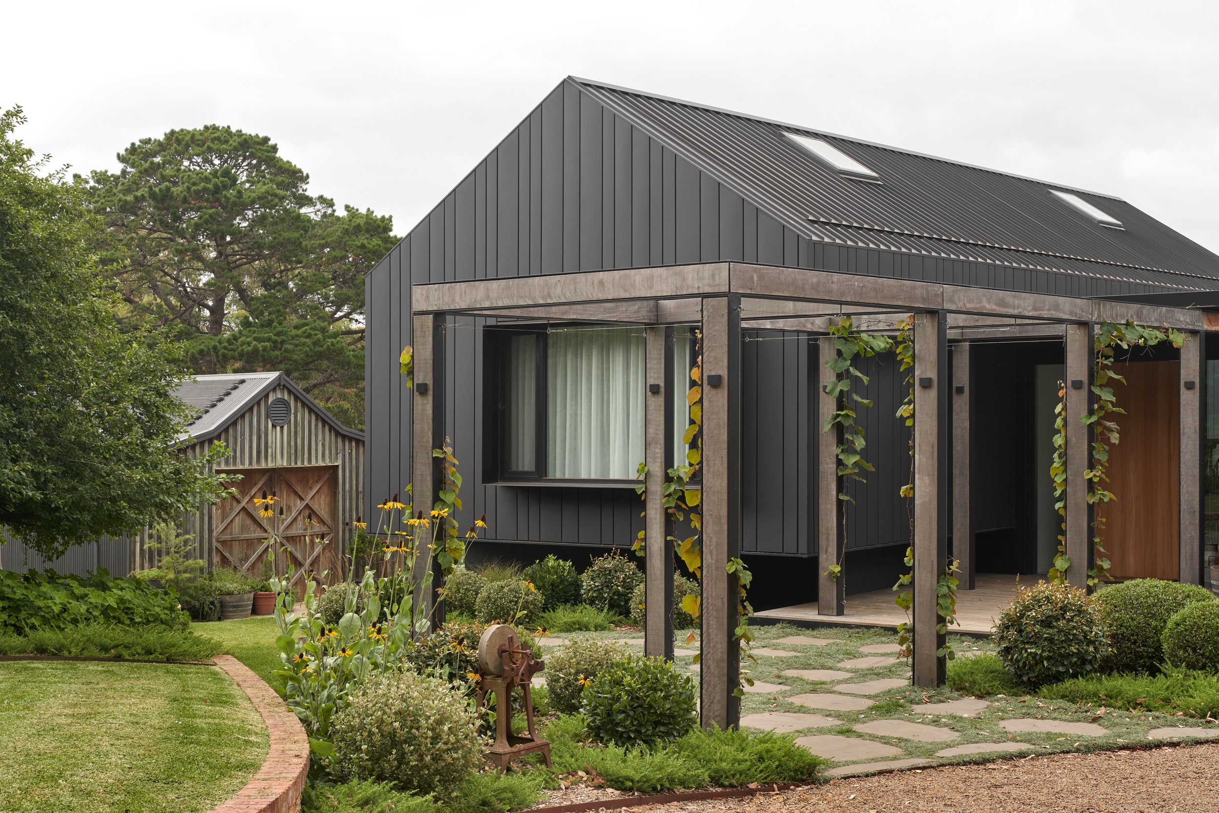 Modern black house with concrete accents and a gabled metal roof, surrounded by a garden with flowers, bushes, and trees.