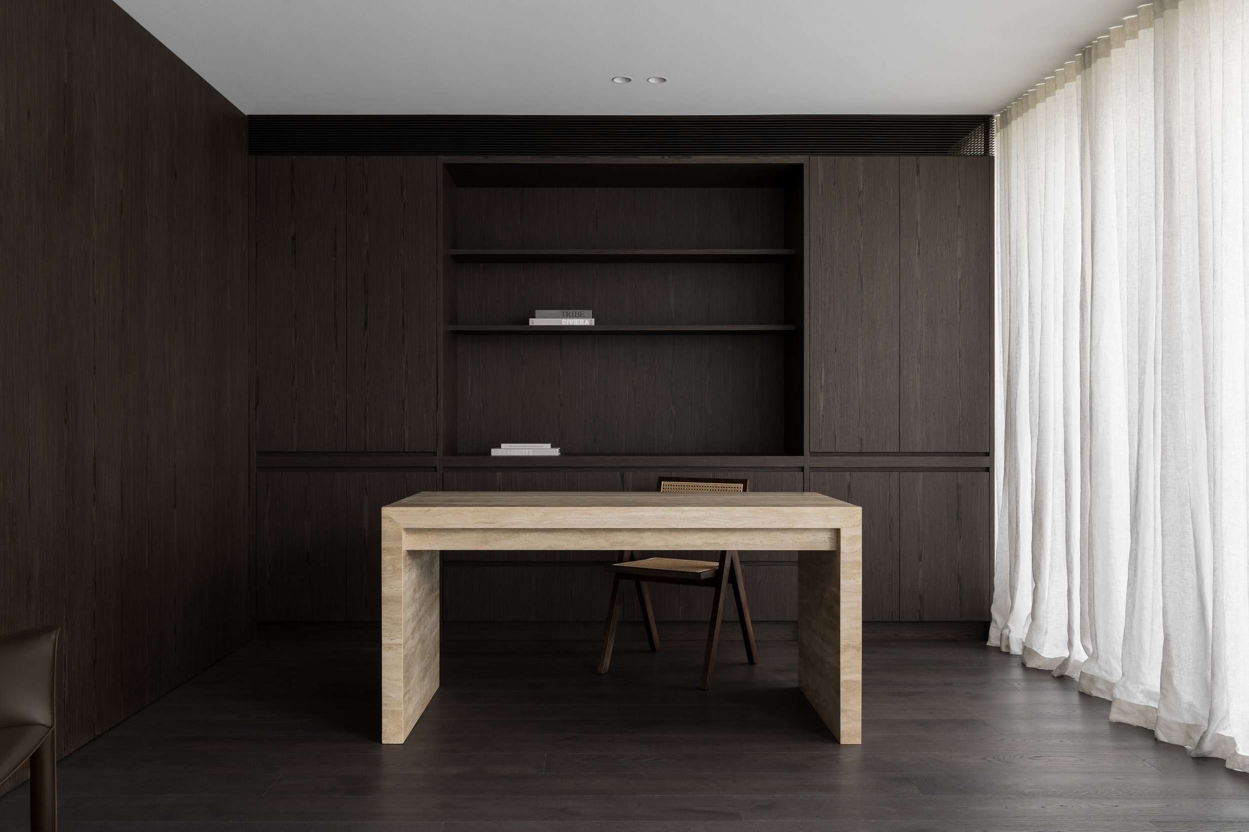 Minimalist office with dark wood-paneled wall, built-in shelving with books, light-colored stone desk, and beige curtains on the right.