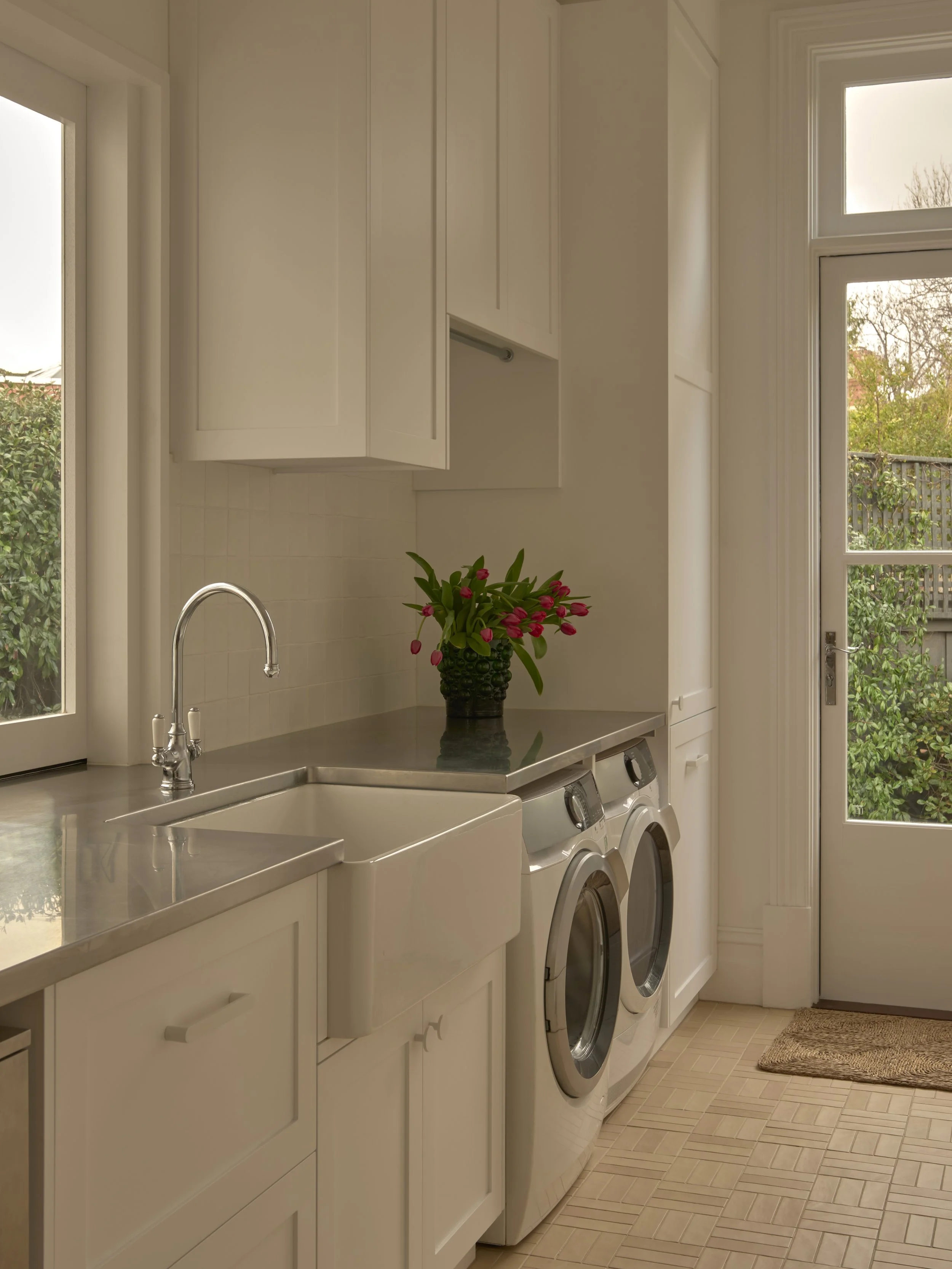 Laundry room with white cabinets, a white utility sink, a front-loading washing machine and dryer side by side, a vase with pink flowers on the countertop, a window and a door leading outside.