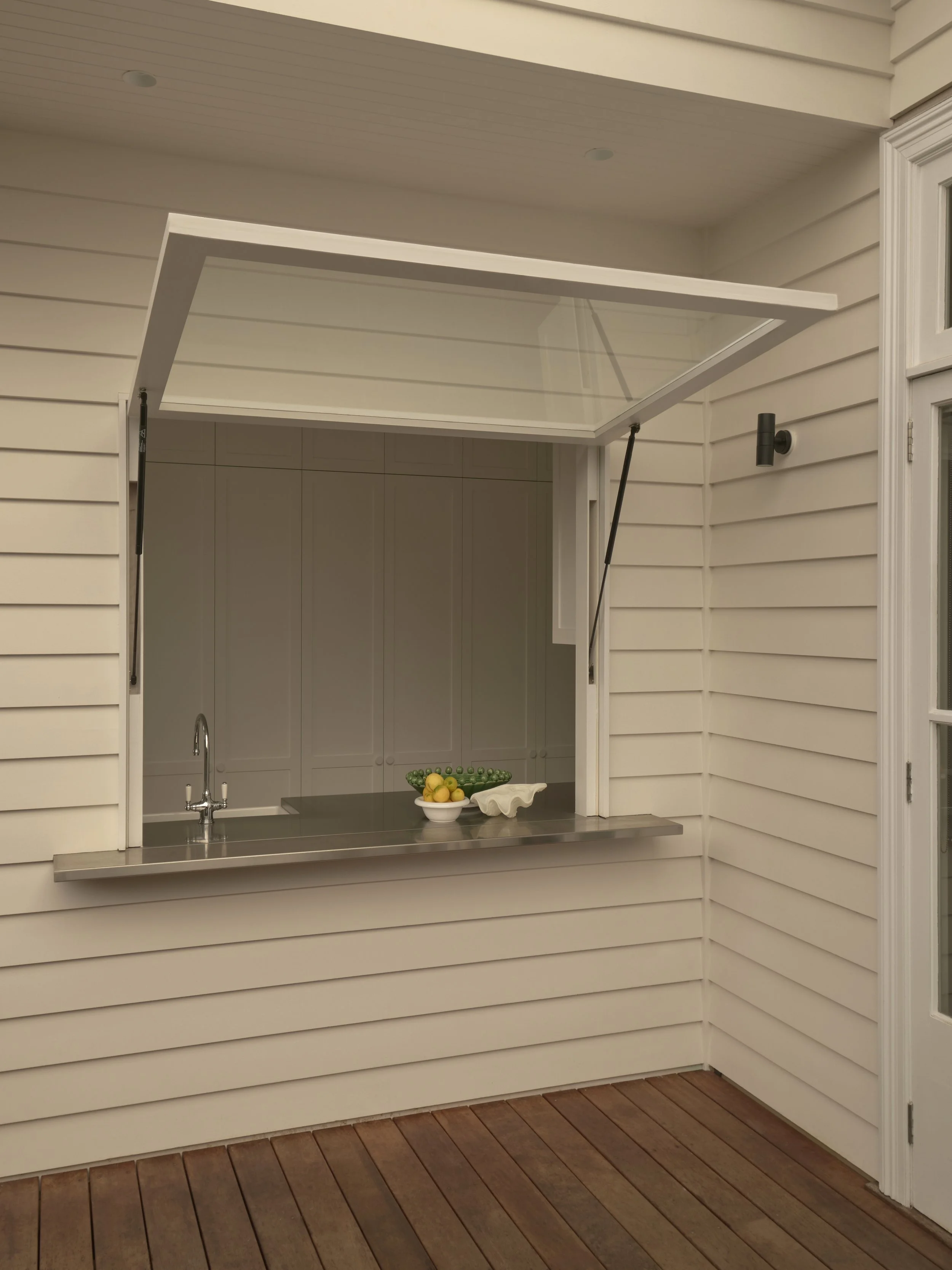 Outdoor kitchen with a countertop and a window opening, open to a room with white cabinets. There are lemons and greens in a bowl on the countertop.