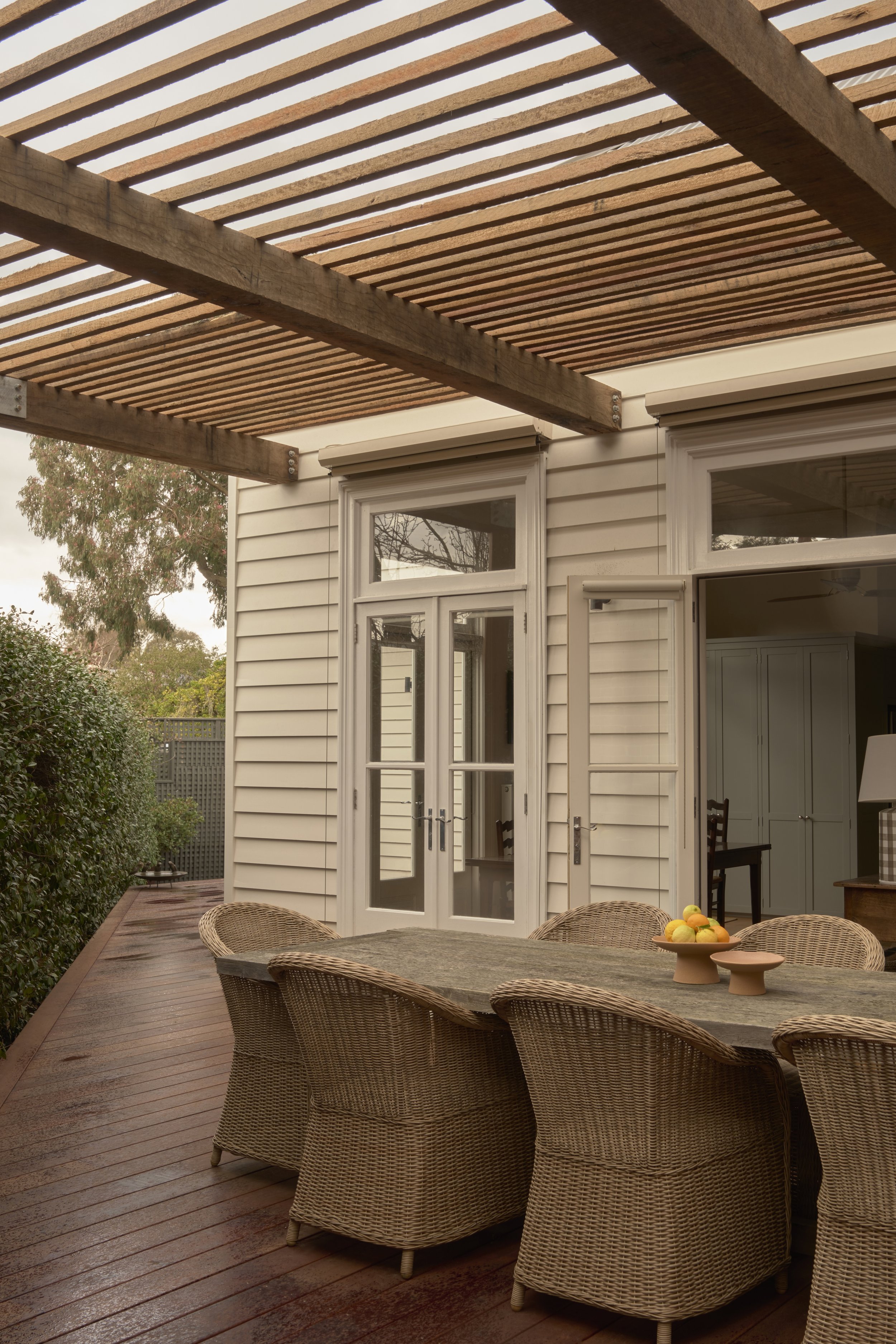 Outdoor patio with a wooden slat pergola, wicker chairs around a rectangular table, and a white house with glass doors.