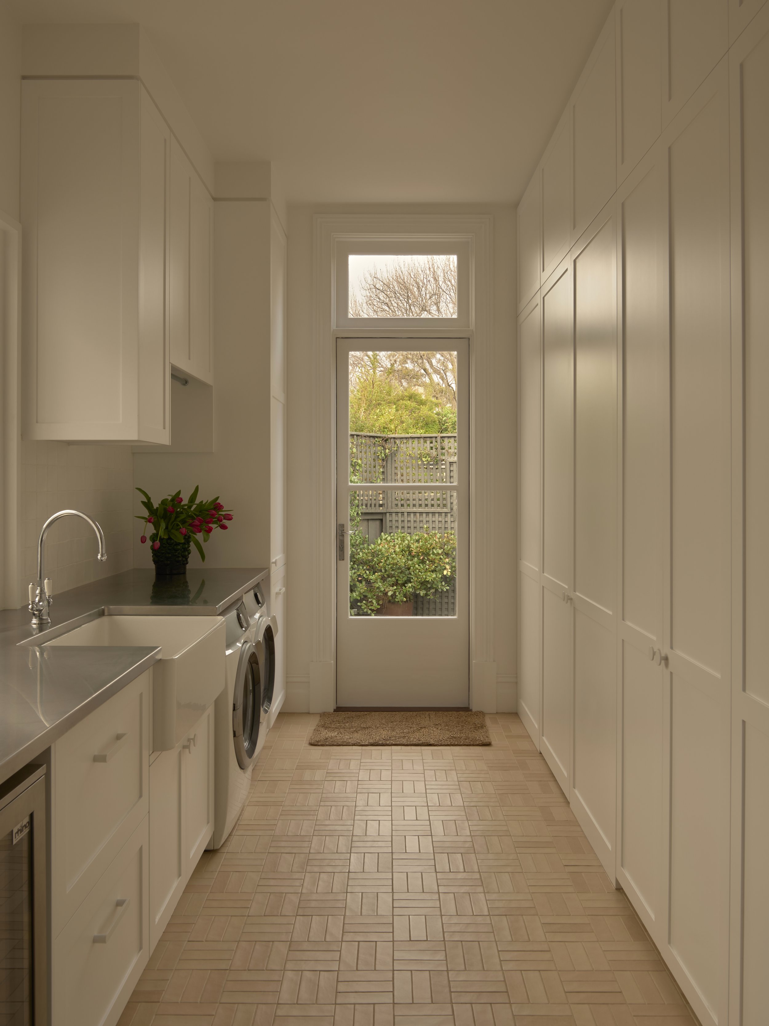 A bright laundry room with white cabinets on both sides, a washing machine and dryer, a stainless steel sink, a potted plant with red flowers, a glass door leading outside, and a view of trees and a garden.