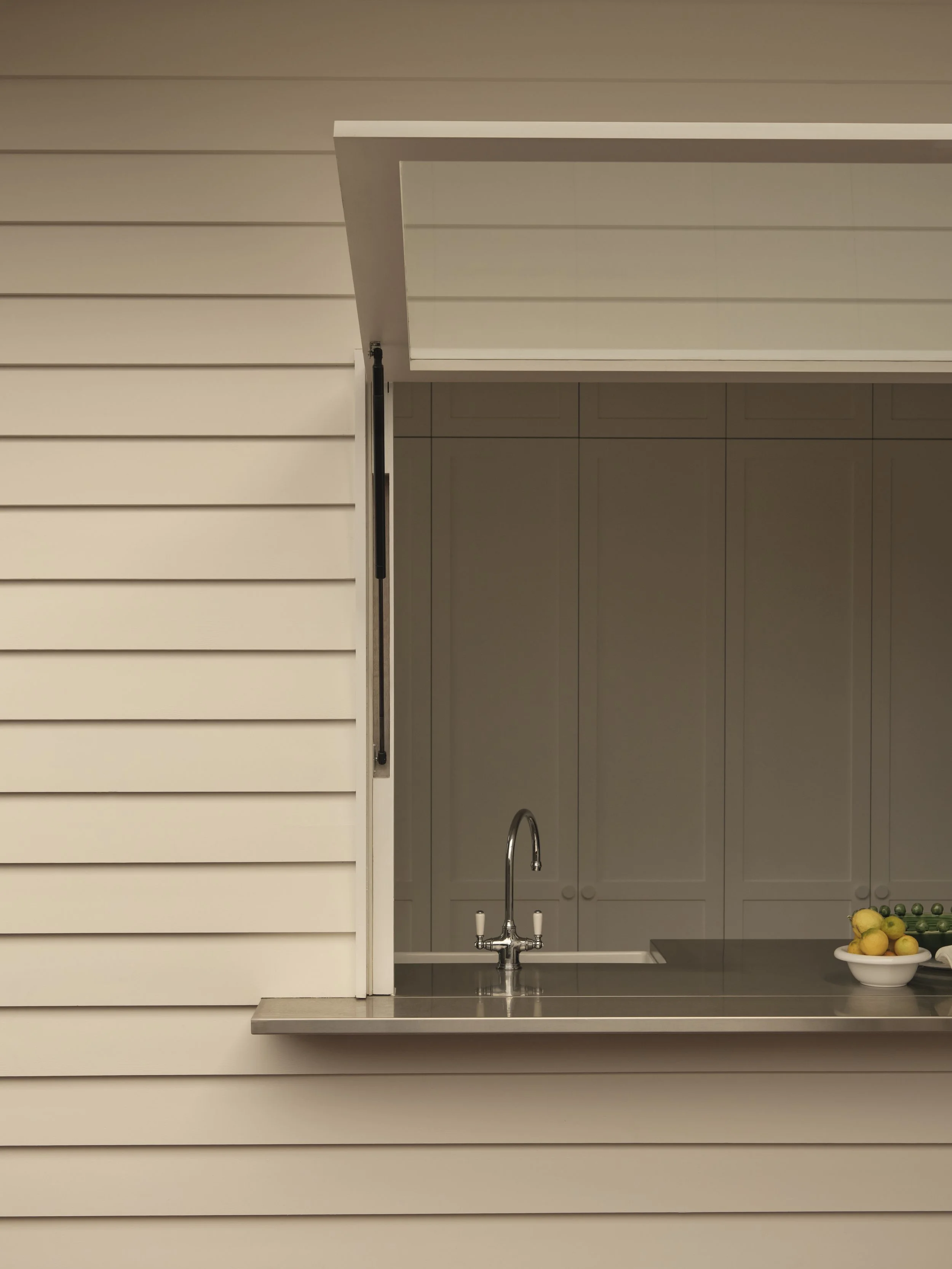 A view of a kitchen through a horizontal sliding window with white siding. The kitchen has white cabinets, a silver faucet, a counter top with a bowl of lemons, and some green peas.