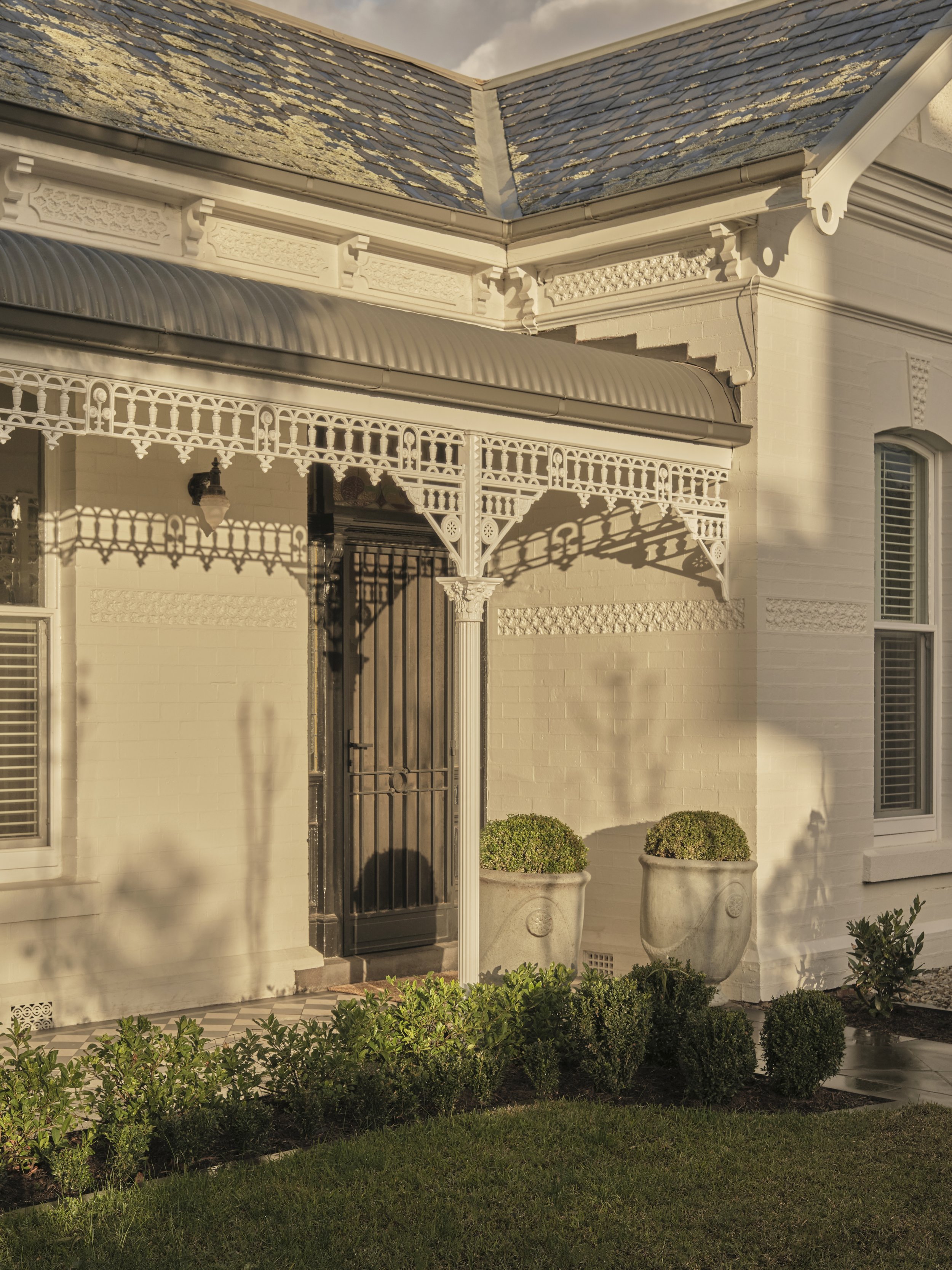 Front porch of a house with ornate white woodwork, a black security door, two large plant pots with bushes, and small shrubs in the garden bed, illuminated by golden sunlight.