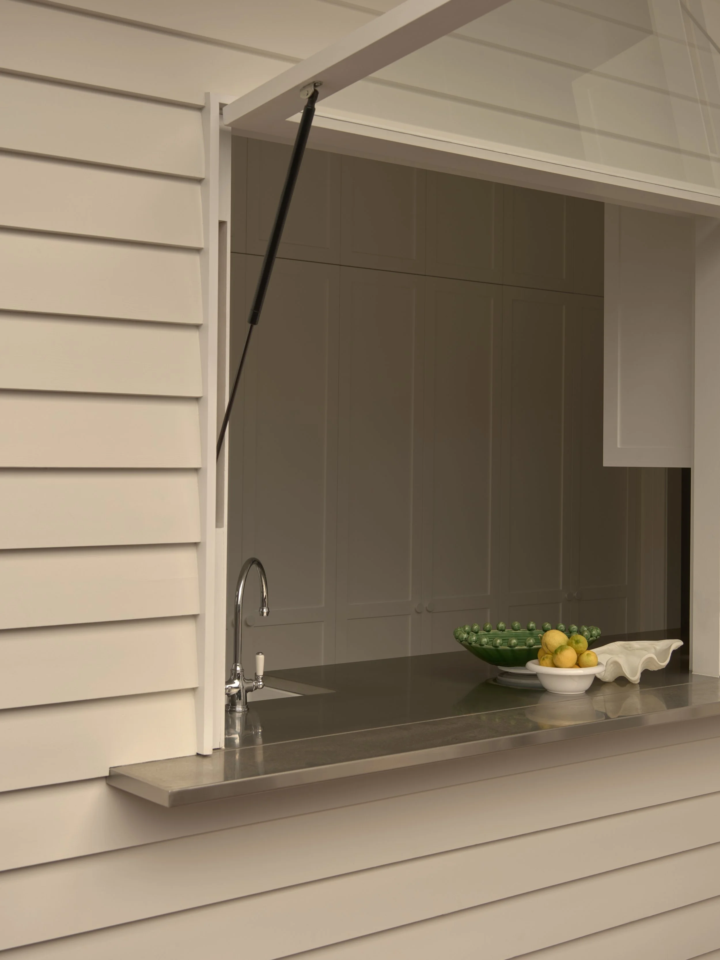 Part of a kitchen window with a metal countertop and ceramic bowls filled with lemons and green grapes, all viewed through a partially open shutter.