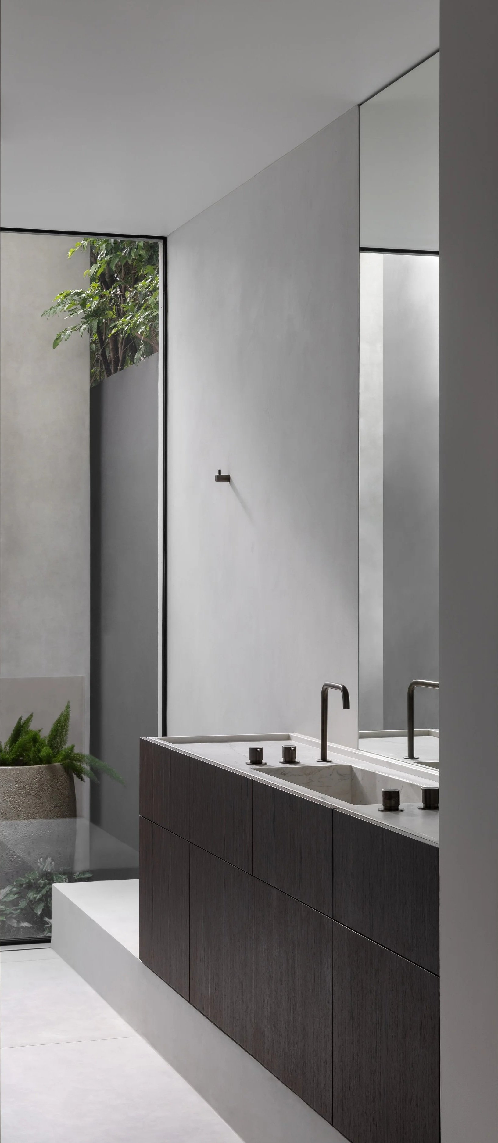 Minimalist bathroom with dark wooden vanity, marble sink, modern black faucet, large mirror, concrete wall, and floor-to-ceiling window with greenery outside.