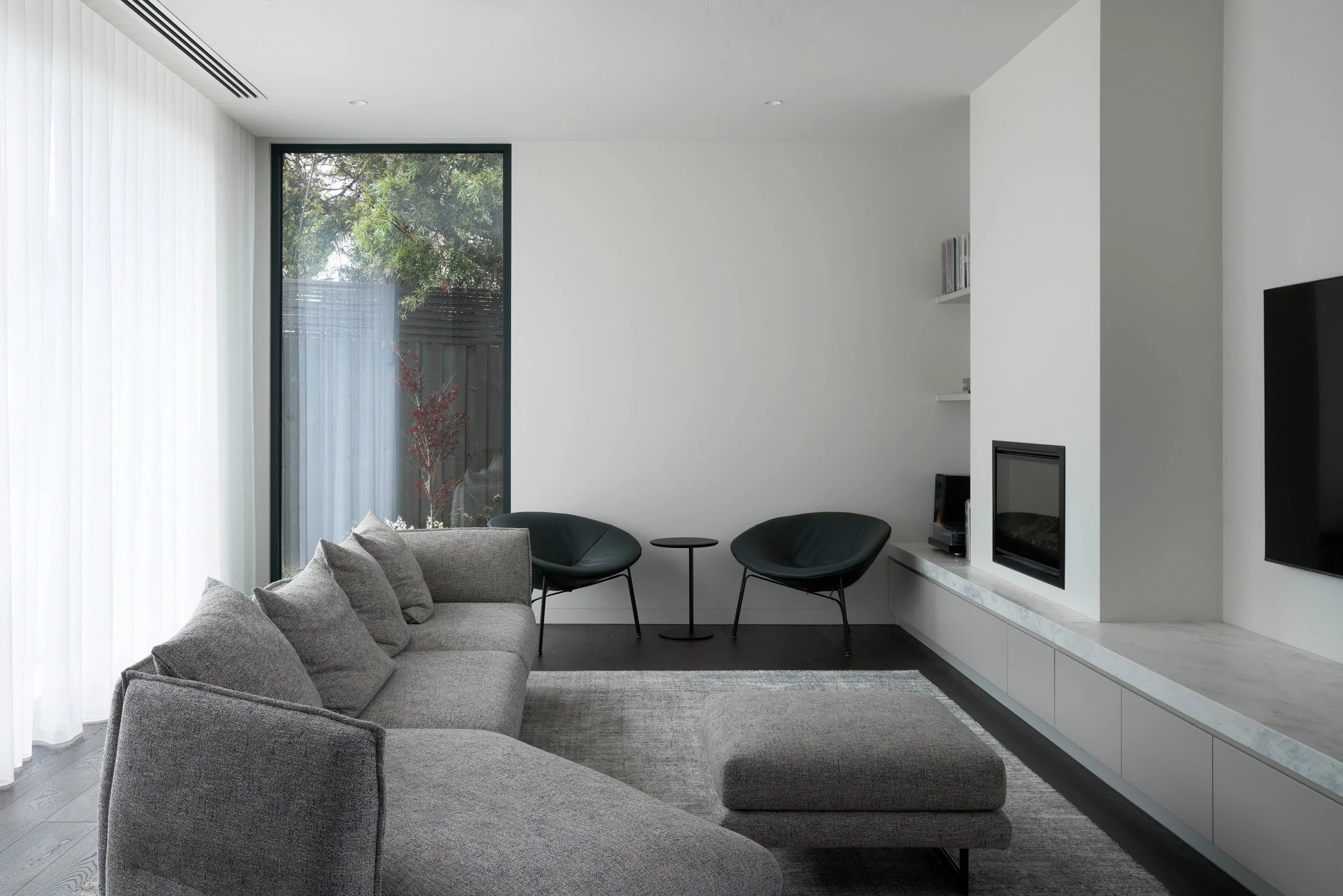A modern living room with a sectional gray sofa, two black lounge chairs, a small round black side table, a white built-in shelf with a fireplace below, a large window with sheer curtains, a gray rug, and a wall-mounted television.