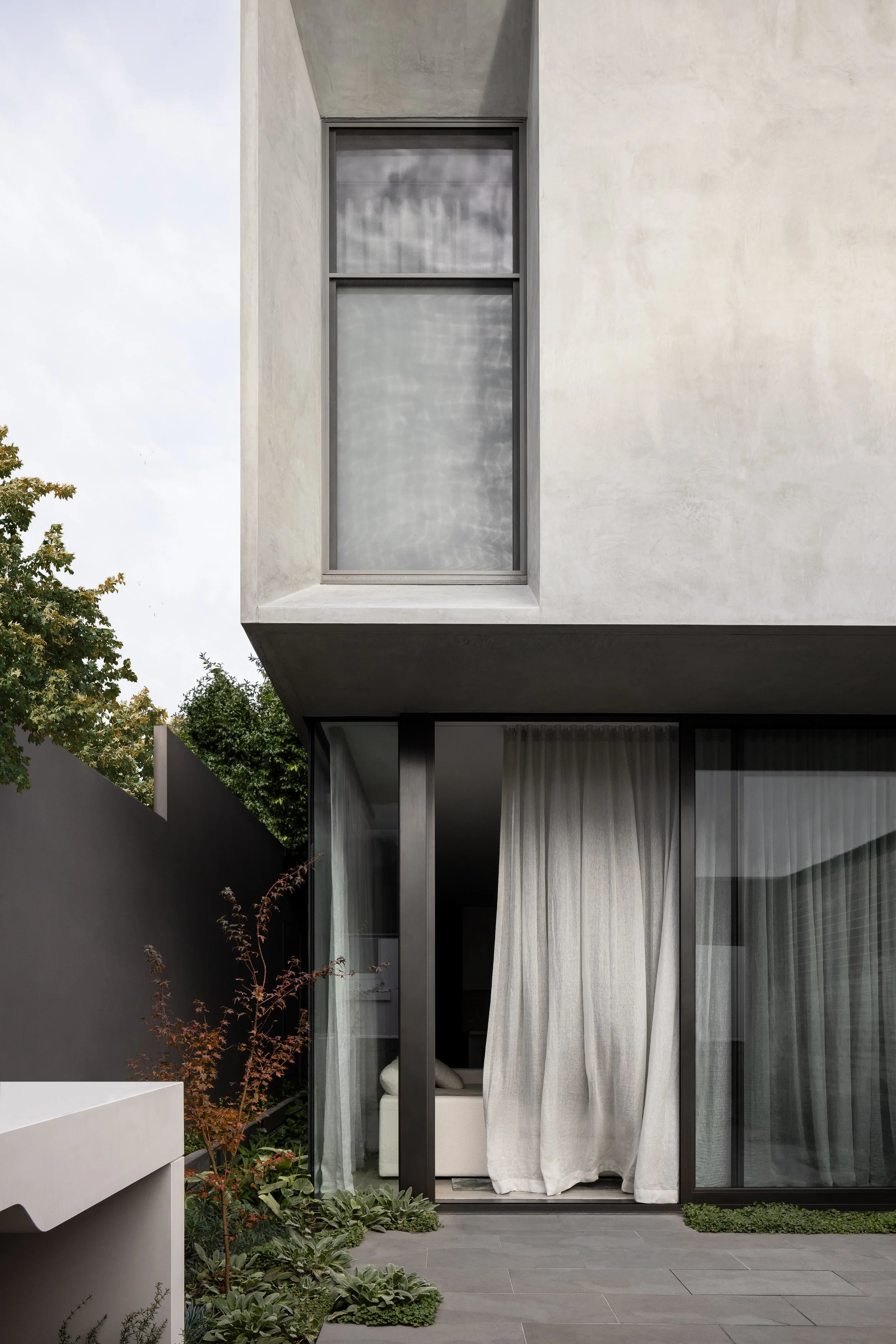 Modern minimalist building with a concrete upper level featuring a large rectangular window and a lower level with large glass sliding doors and white curtains, surrounded by a small garden and patio.