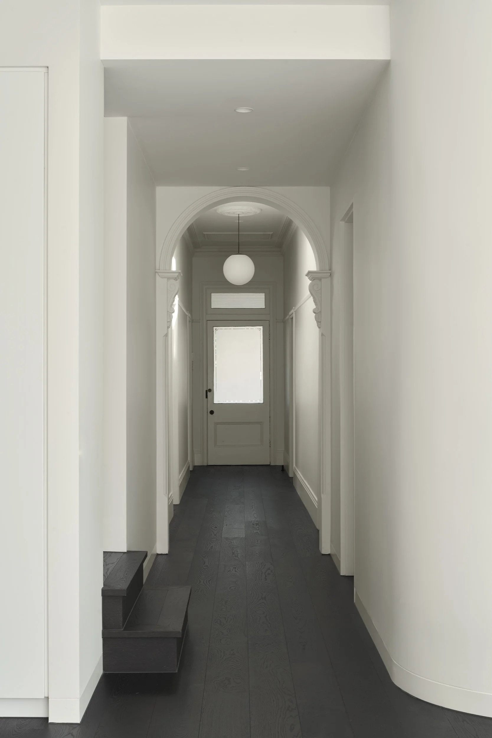 White hallway with dark wood flooring, an arched doorway, and a door at the end with frosted glass, illuminated by a hanging globe light.