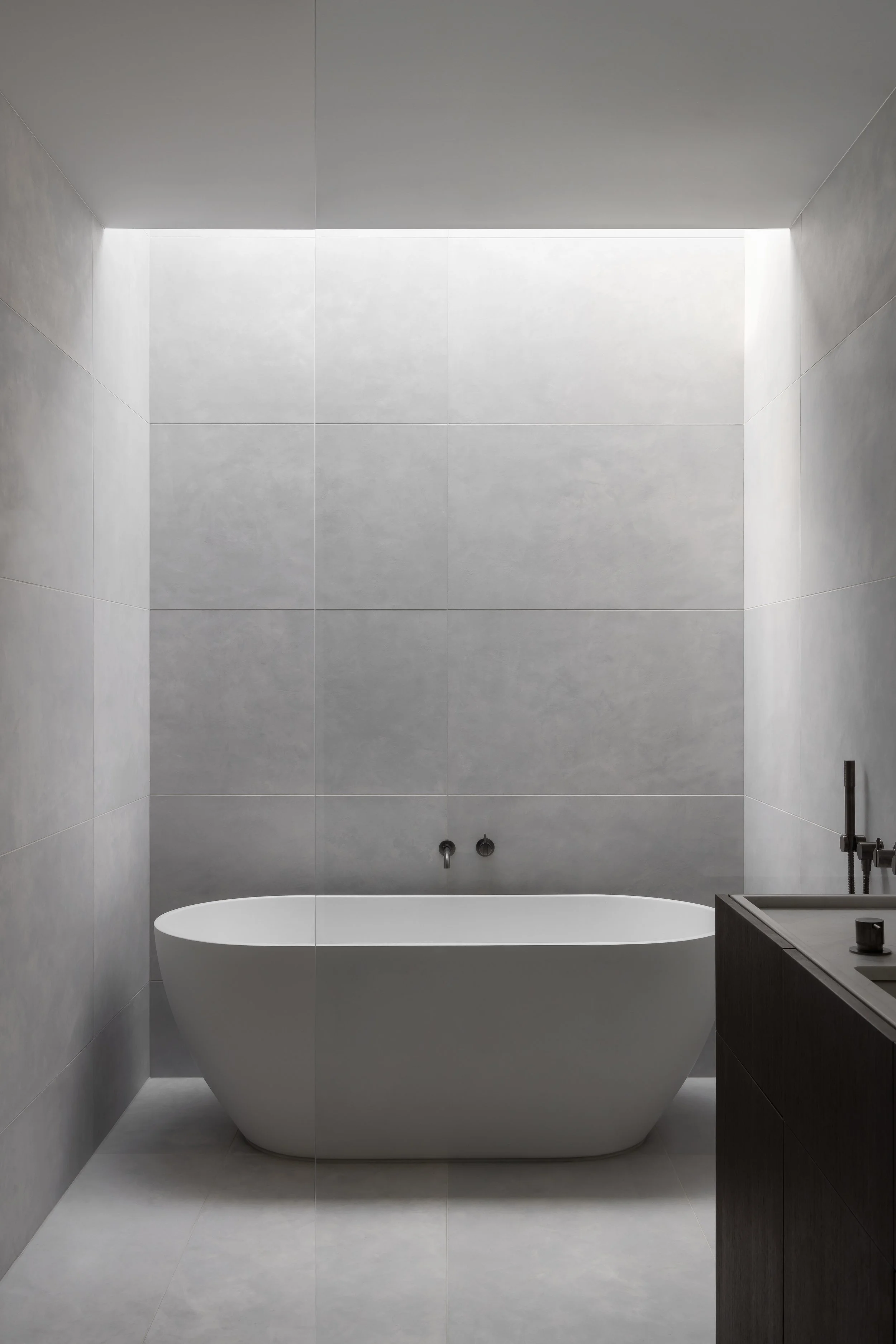 Modern bathroom with a freestanding white bathtub, gray tiled walls, and a dark vanity with a sink on the right side.