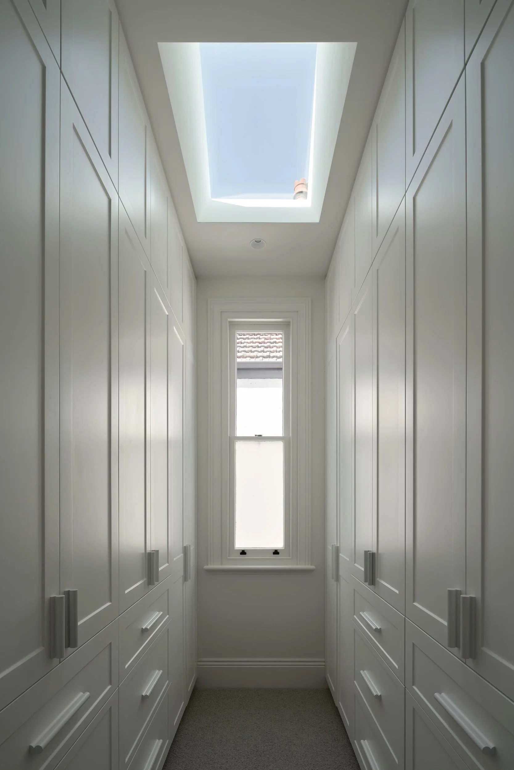A bright, white walk-in wardrobe with built-in cabinets on both sides, a small window at the end, and a skylight overhead.