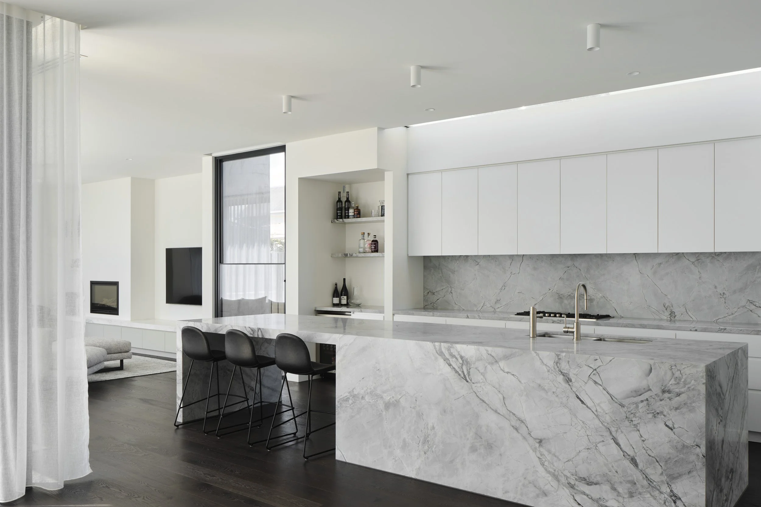 Modern kitchen with white cabinets and marble countertops, bar stools, and a window with sheer curtains.
