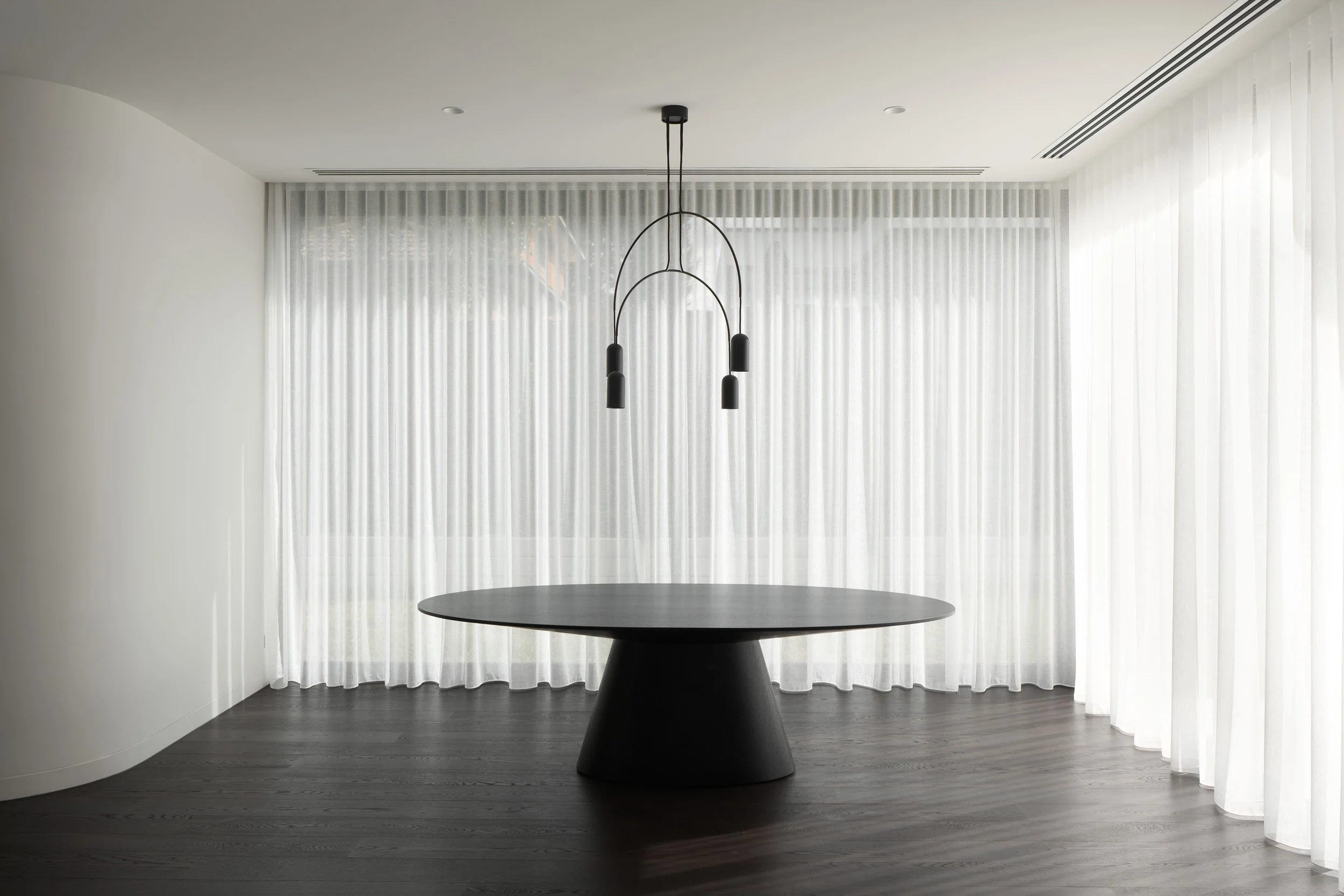 Minimalistic dining room with dark round table, black pendant light, white curtains, and dark hardwood floor.